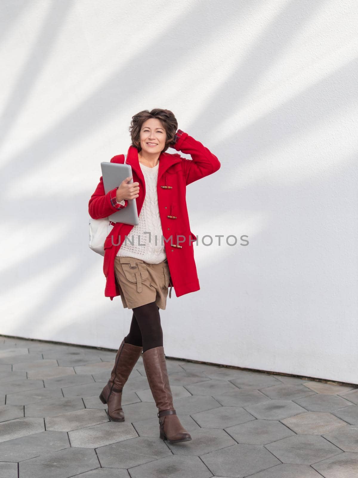 Curly woman in red duffle coat is walking by white wall. Smiling student with laptop. Freelancer with portable device for work. Modern lifestyle.