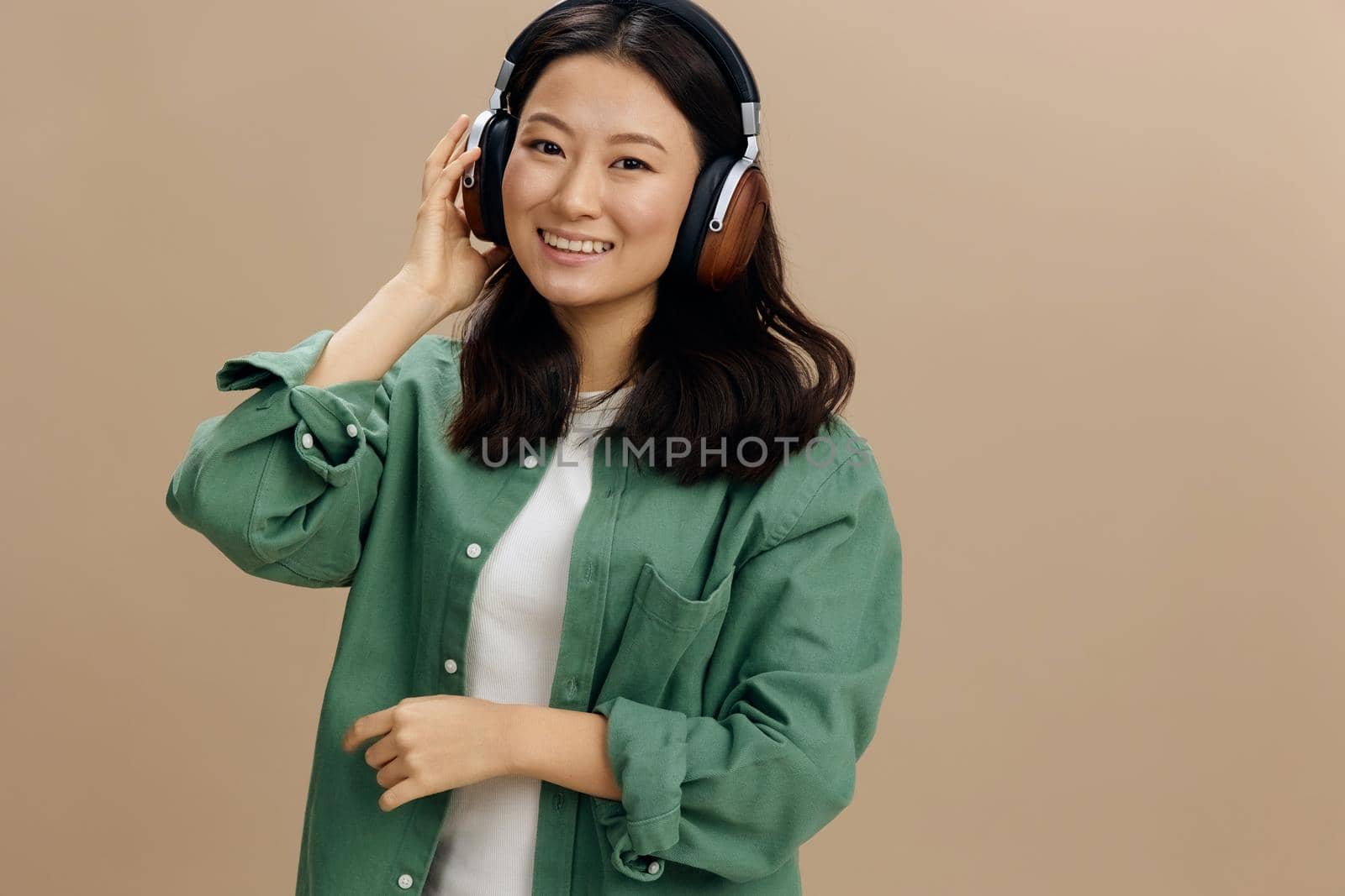 Enjoy this sound. Happy cute Asian student young woman in khaki green shirt touch headphones posing isolated on over beige pastel studio background. Cool fashion offer. Music App Platform Ad concept