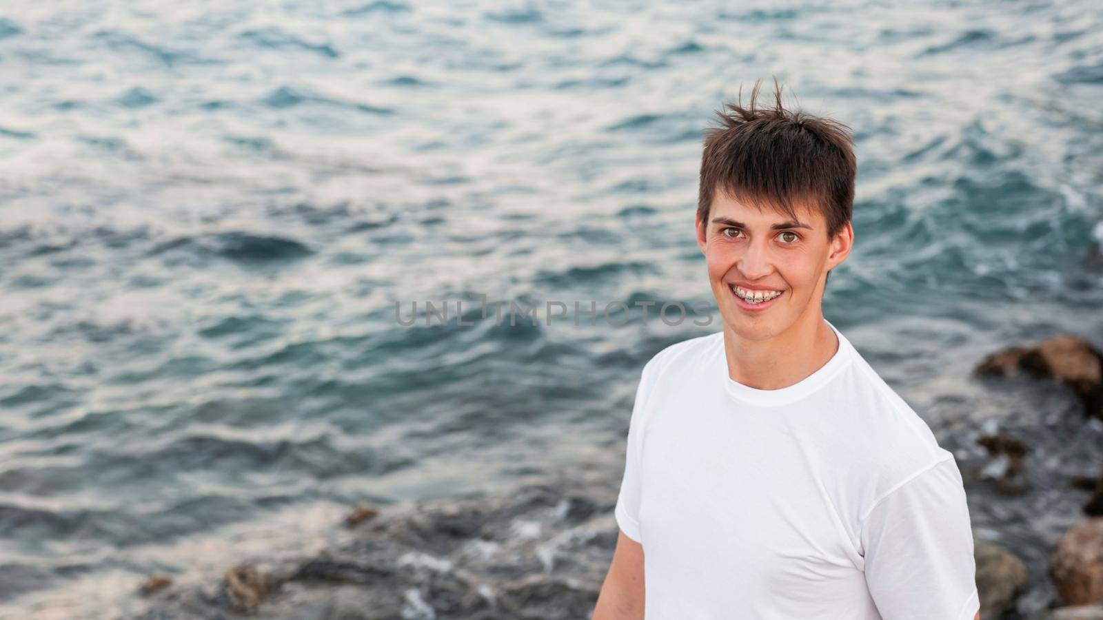 Smiling young man with braces on teeth. Caucasian man looks happily on blue sea background. Horizontal banner with copy space. by aksenovko