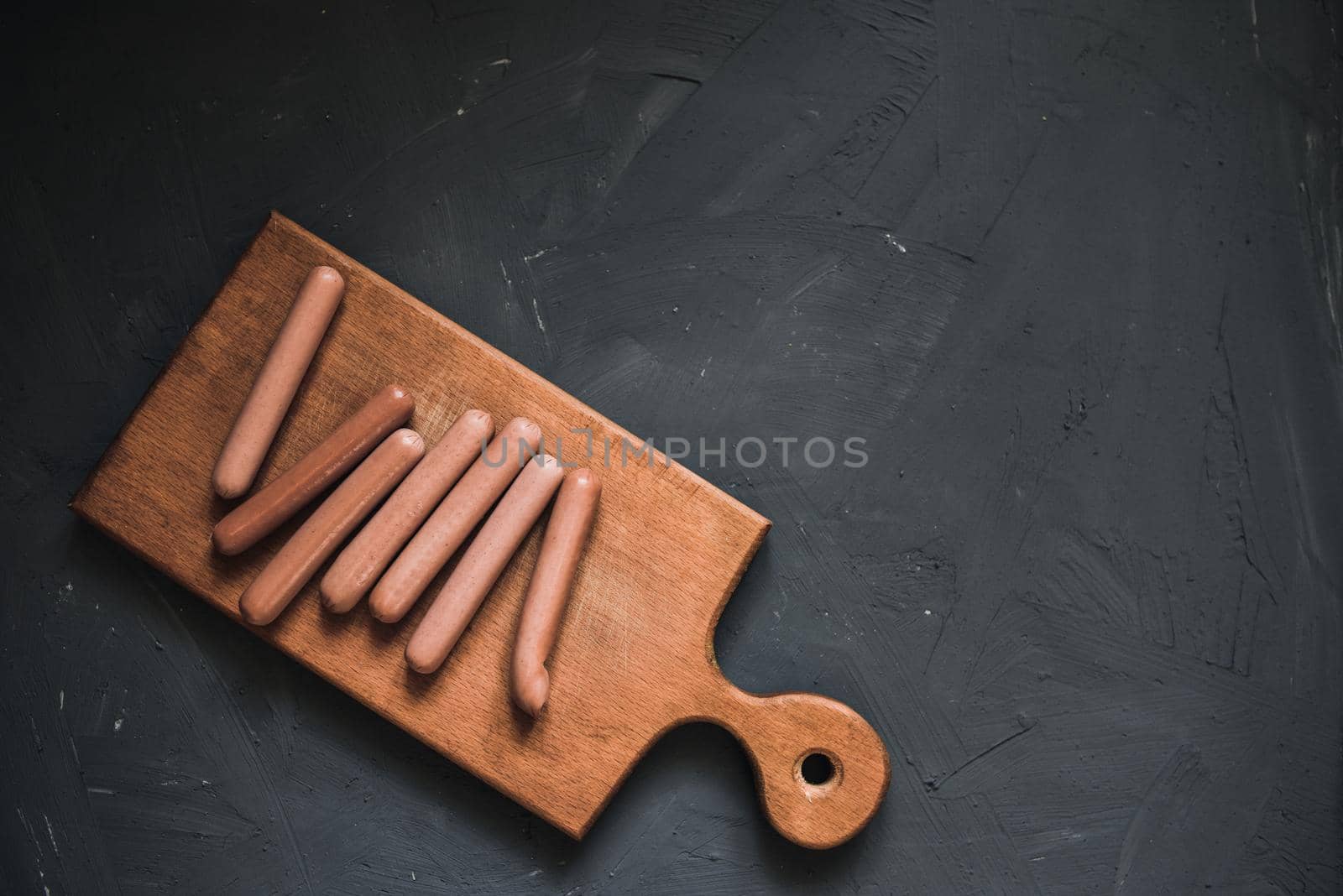 Boiled fried sausages sausages lie on a wooden kitchen board scratched against a dark concrete background.