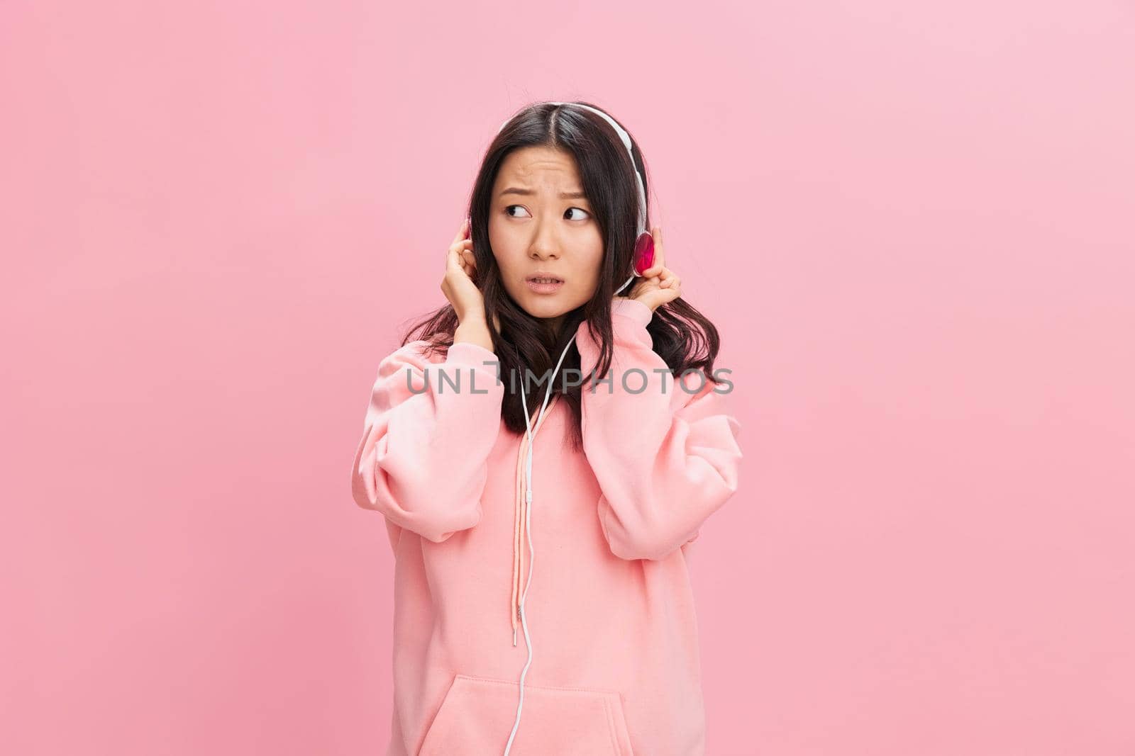 Closeup portrait of shocked cute Asian student young lady in pink hoodie sweatshirt with cute headphones posing isolated on over pink studio background. Good offer. Sound streaming platform ad concept
