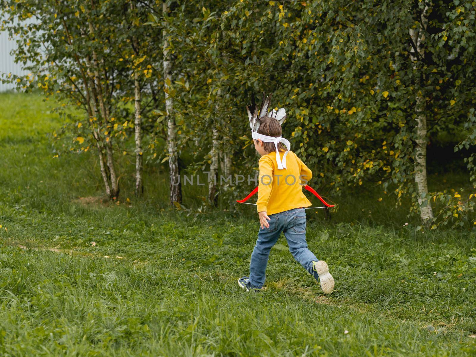 Little boy is playing American Indian on field. Kid has handmade headdress made of feathers and bow with arrows. Costume role play. Outdoor leisure activity. Fall season.