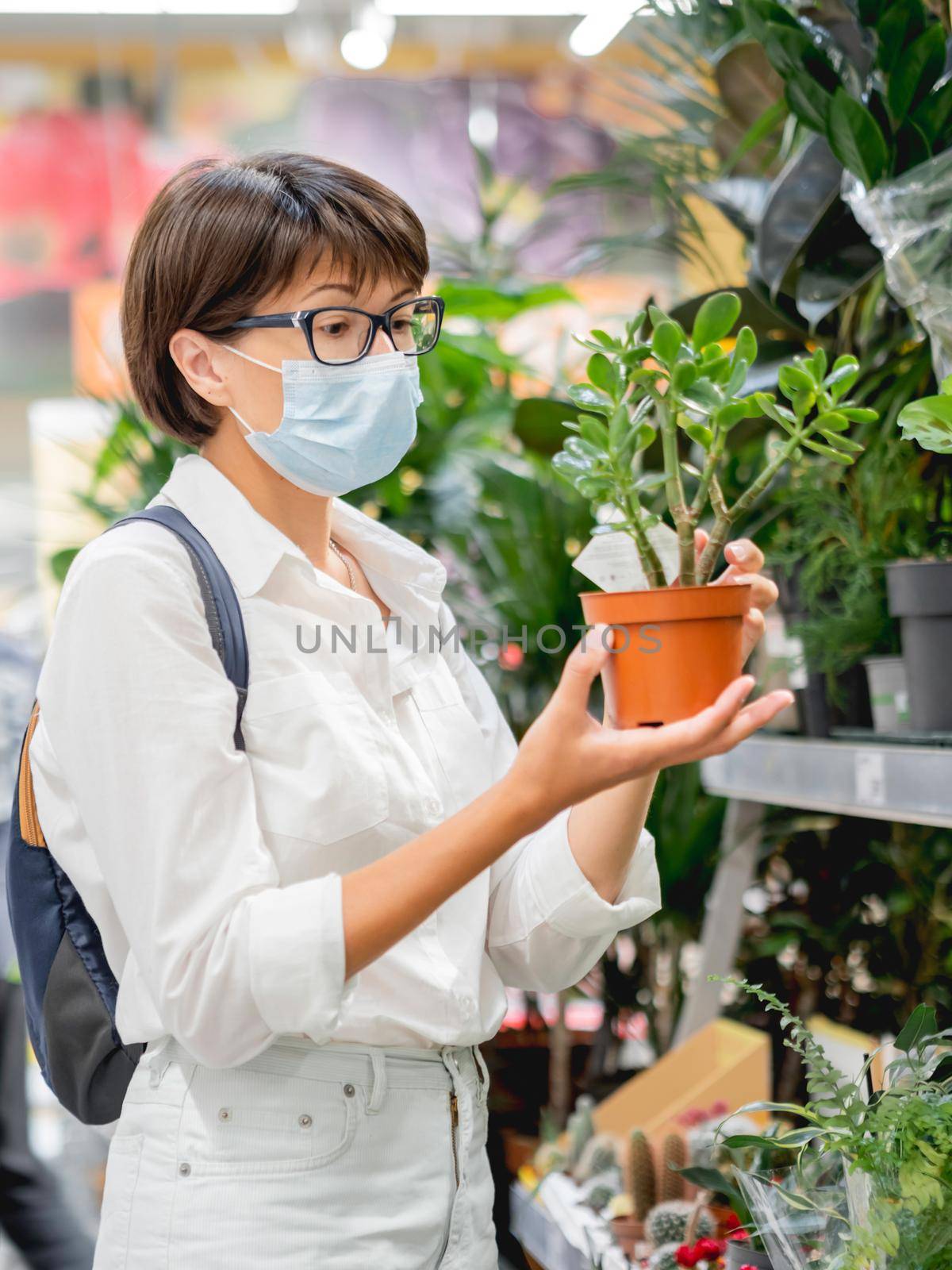 Woman in medical protective mask chooses plants for home. Shelves with seedlings, flowering plants and seeds in flower shop. Indoor agronomic market. by aksenovko
