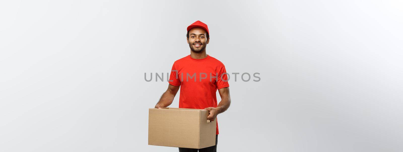 Delivery Concept - Portrait of Happy African American delivery man in red cloth holding a box package. Isolated on Grey studio Background. Copy Space. by Benzoix