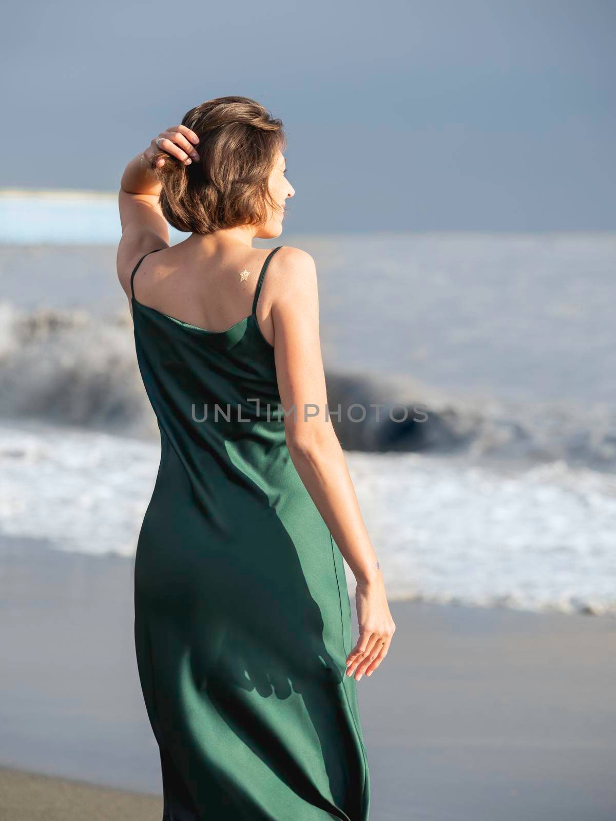 Caucasian woman in green silk dress. Pretty woman at seaside. Long-awaited vacation. Golden star temporary tattoo. View from the back.