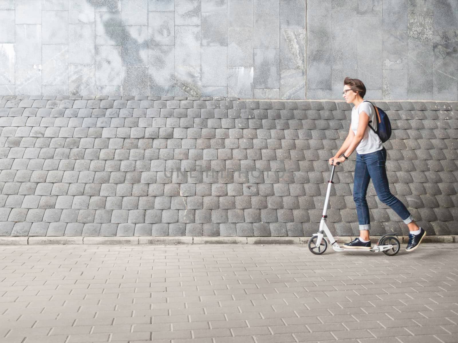 Woman rides kick scooter. Female in jeans and sneakers moves fast on grey tiled wall background. Healthy lifestyle. Eco-friendly urban transport. by aksenovko