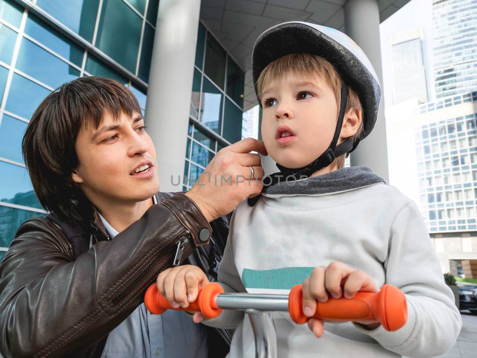 Father is fastening belt of his son's helmet. Kid is training to ride kick scooter. Safety first. Moral support. Urban lifestyle.