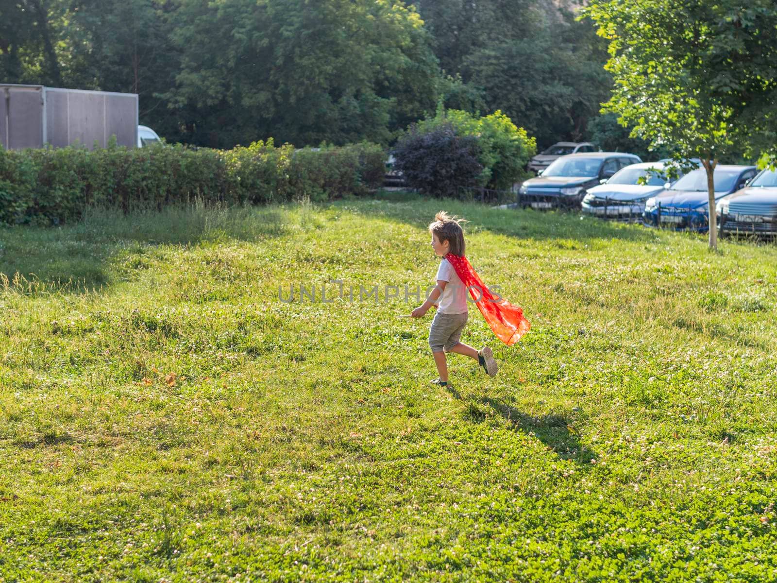 Little boy is playing superhero on lawn. Kid in handmade bright red cloak. Outdoor role-playing game. Costume play at sunlight.
