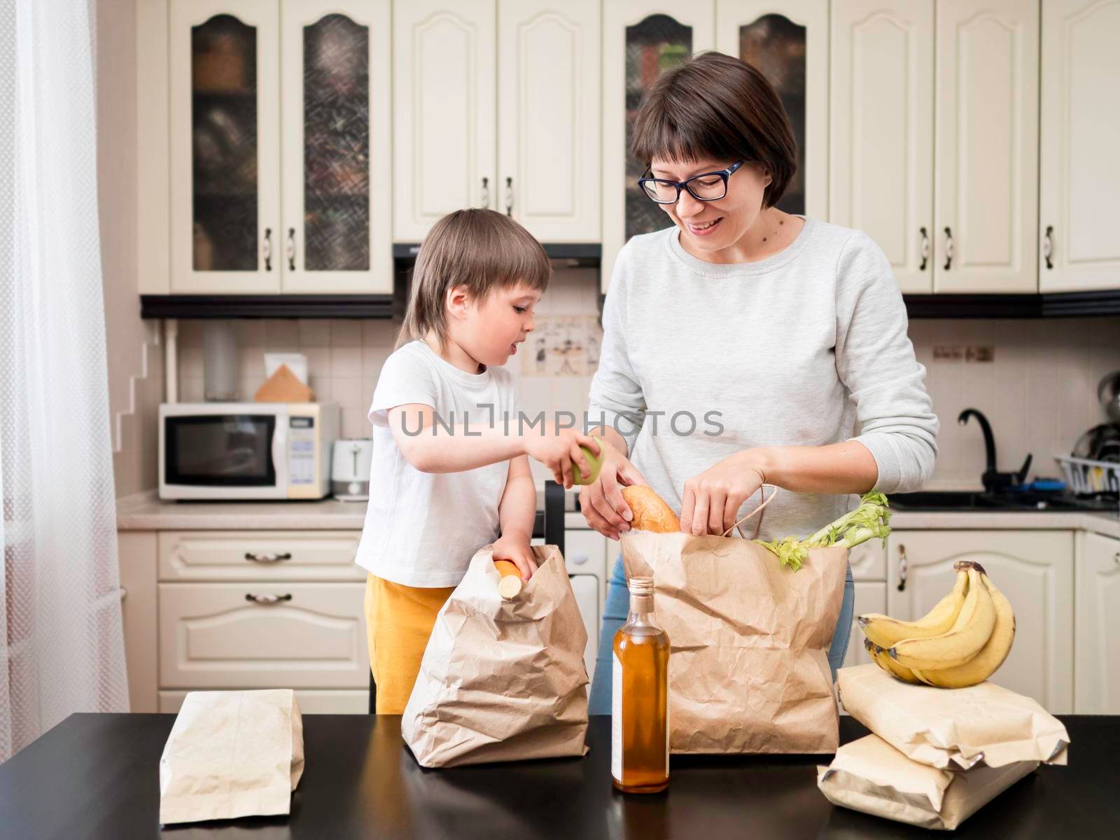 Woman and toddler boy sorts out purchases in the kitchen. Mother and son with grocery delivery in paper bags. Subscription service from grocery store. by aksenovko