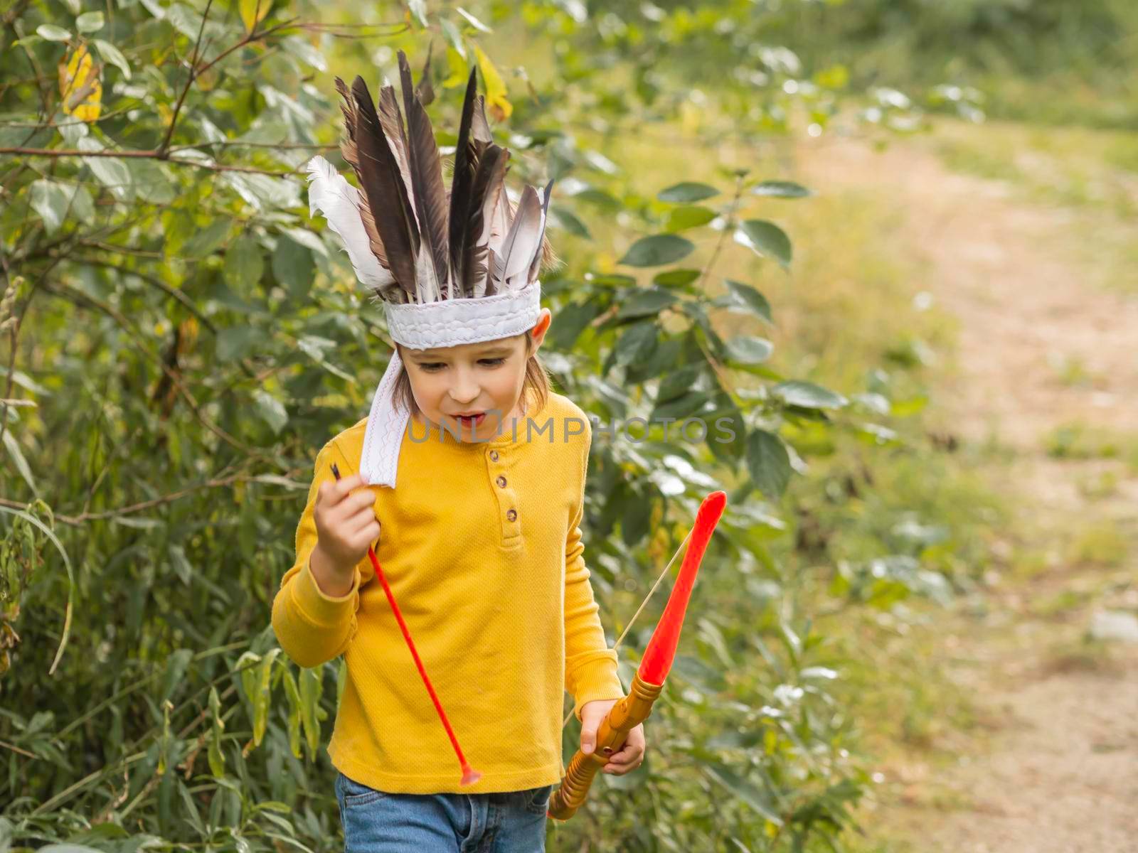 Little boy is playing American Indian on field. Kid has handmade headdress made of feathers and bow with arrows. Costume role play. Outdoor leisure activity. Fall season. by aksenovko