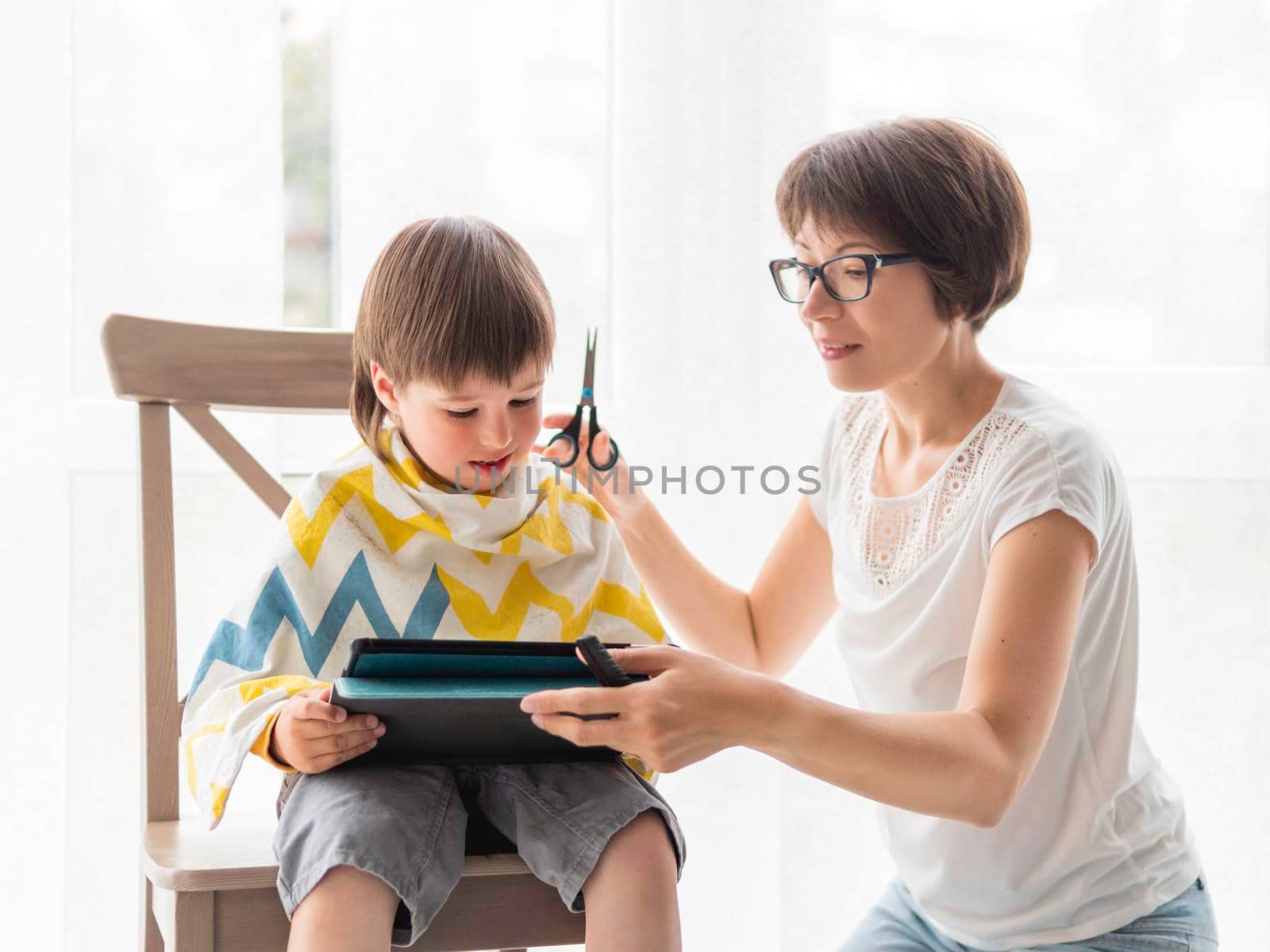 Mother cuts her son's hair by herself. Little boy sits with digital tablet. New normal in case of coronavirus COVID-19 quarantine and lockdown.