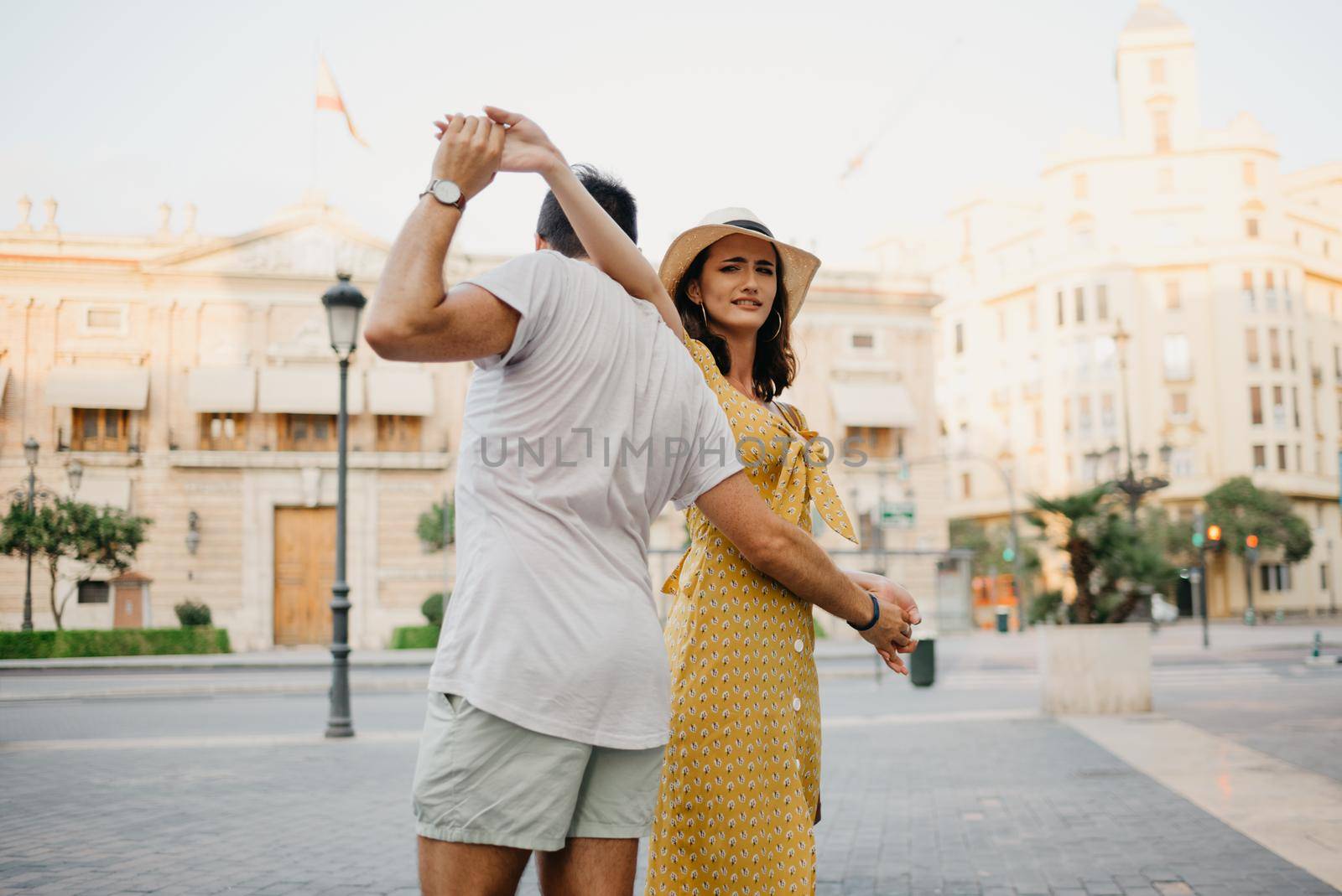 A happy girl in a hat and a yellow dress with a plunging neckline is dancing with her boyfriend with a beard and sunglasses in the old town. A couple of tourists on the sunset in Valencia.