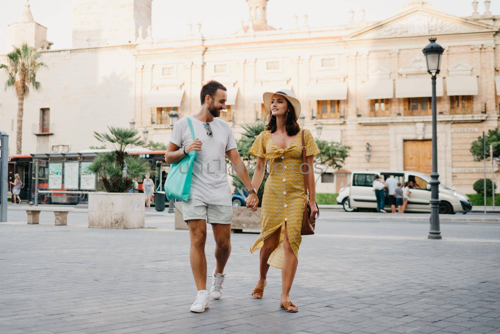 Girl in a hat and a dress with a plunging neckline and her boyfriend with a beard and sunglasses are relaxing holding each other's hand in the old town. A couple of tourists on the sunset in Valencia.