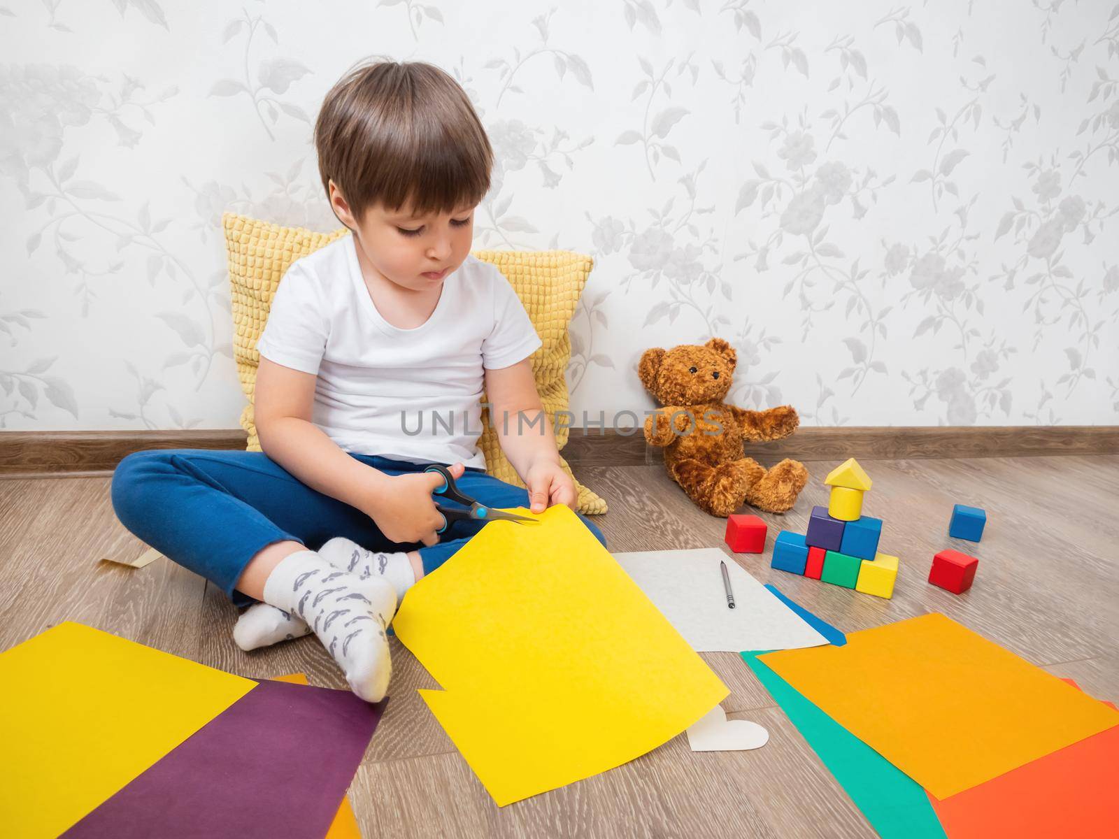 Toddler boy learns to cut colored paper with scissors. Kid sits on floor in kids room with toy blocks and teddy bear. Educational classes for children. Developing feeling sensations and fine motor skills at home. by aksenovko