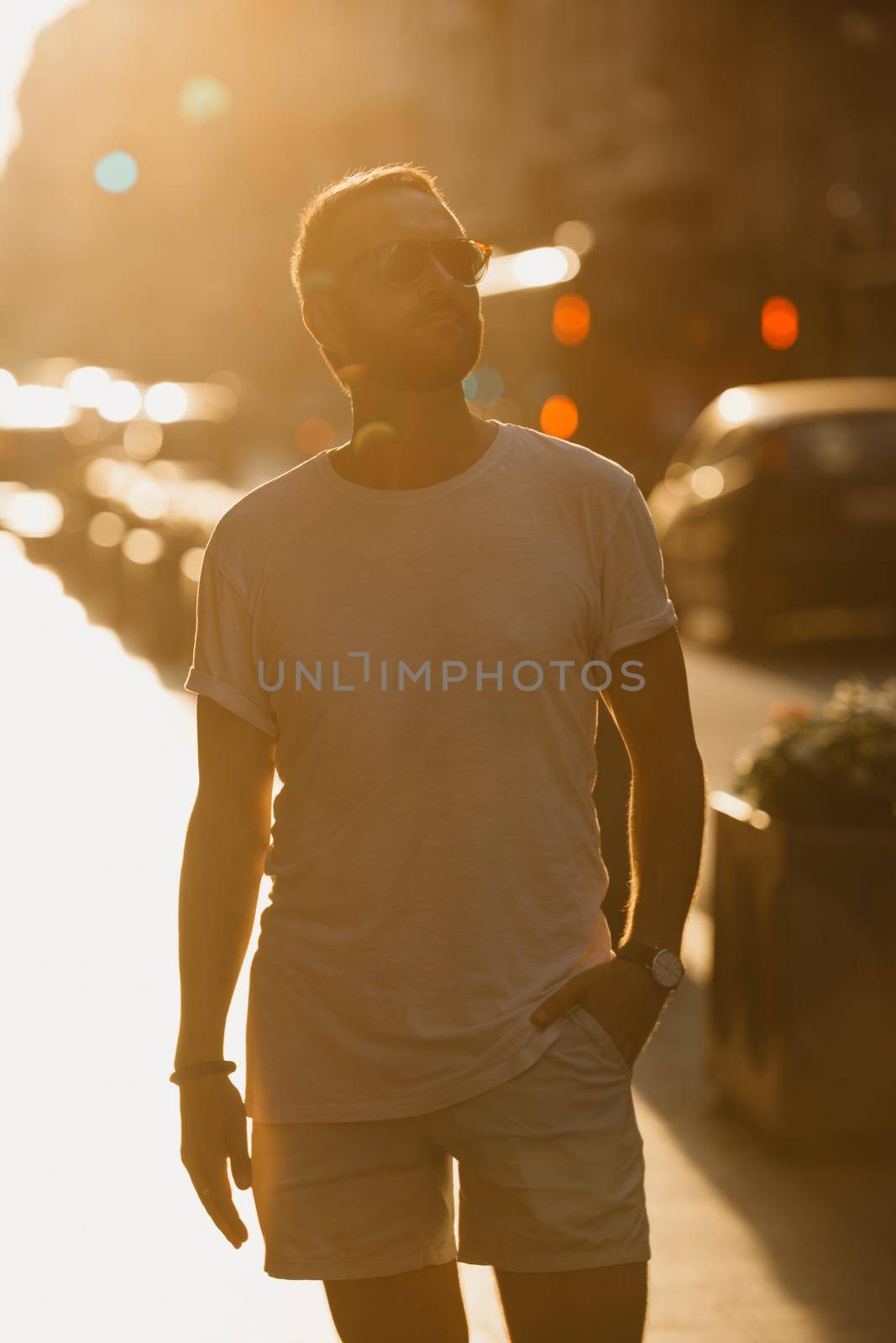 A man in sunglasses, a white t-shirt, and shorts is sightseeing of Valencia in rays of the sunset on an old street. A guy with a beard is enjoying Spain in the evening.