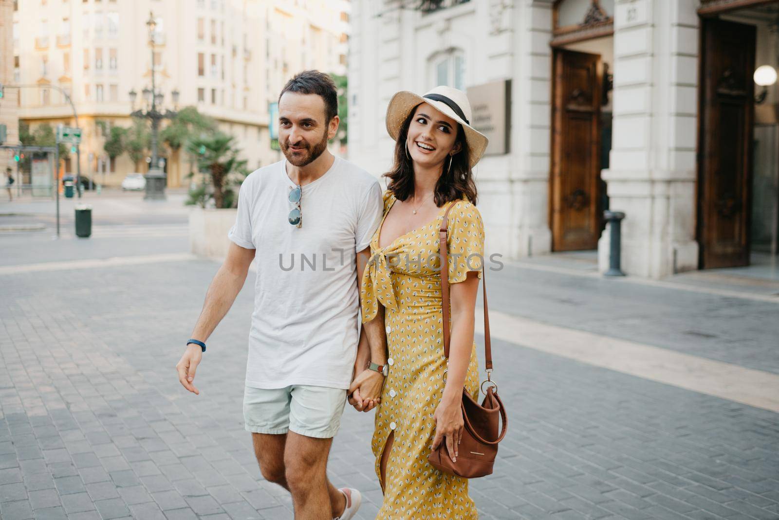 A happy girl in a hat and a yellow dress with a plunging neckline and her boyfriend with a beard and sunglasses are walking holding each other's hand in the old town. A couple of tourists in Valencia.