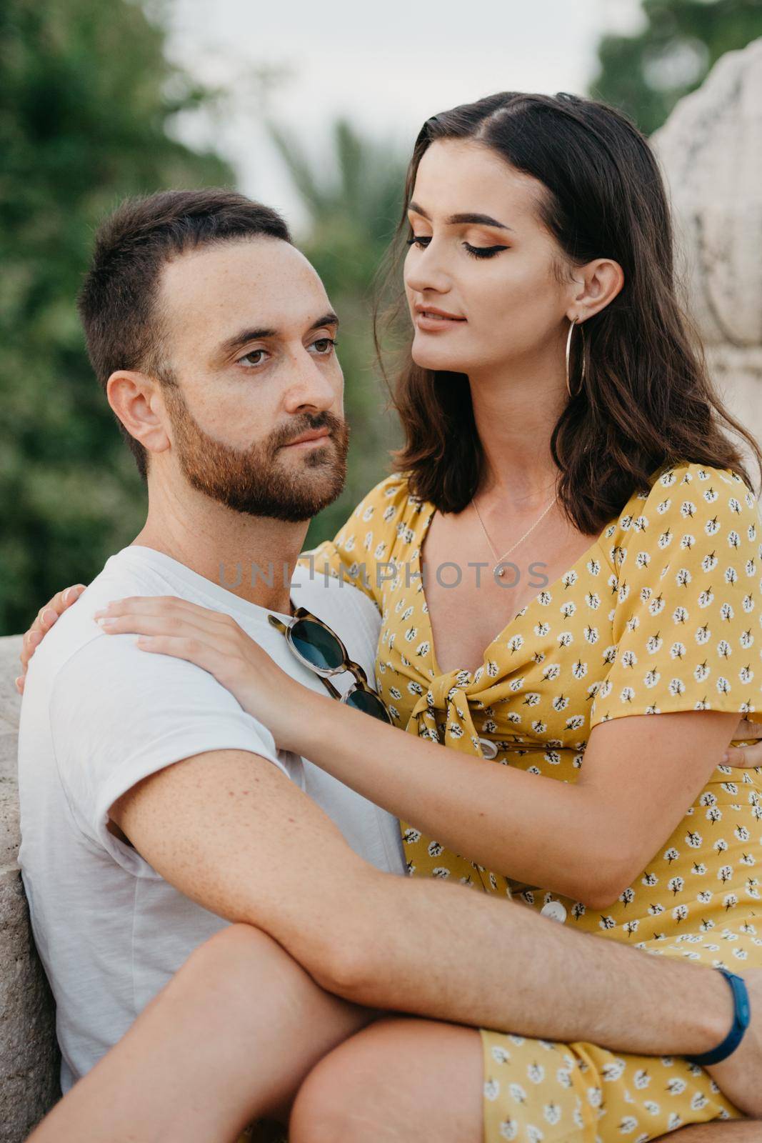 A close photo of a brunette girl in a yellow dress who is sitting on the legs of her boyfriend with a beard on the ancient bridge in old Spain town. A couple of tourists on a date in Valencia.