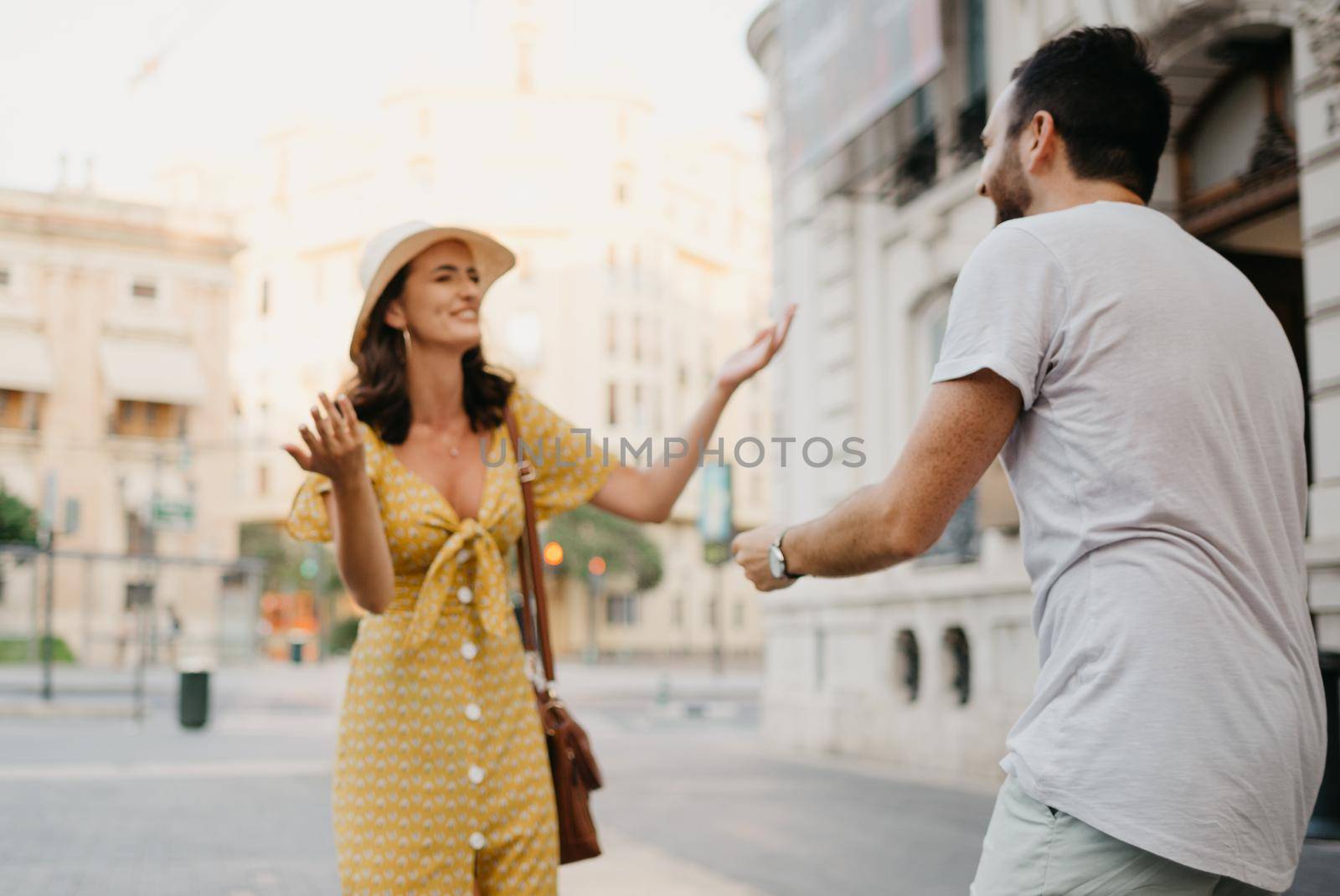 A laughing girl in a hat and a yellow dress with a plunging neckline is joking with her boyfriend with a beard and sunglasses in the old town. A couple of tourists on the sunset in Valencia.