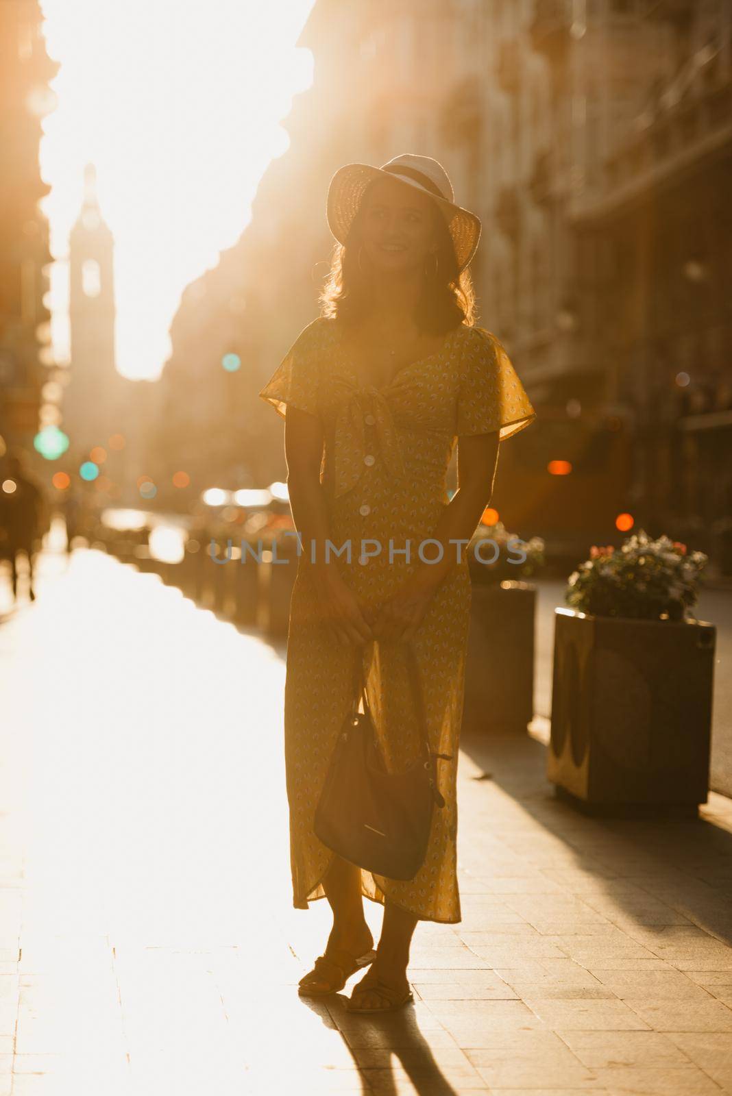 A cute young woman in a hat and a yellow dress with a plunging neckline is holding a leather bag on the sunset in Spain. A gorgeous smiling girl is posing on an old street in Valencia in the evening.
