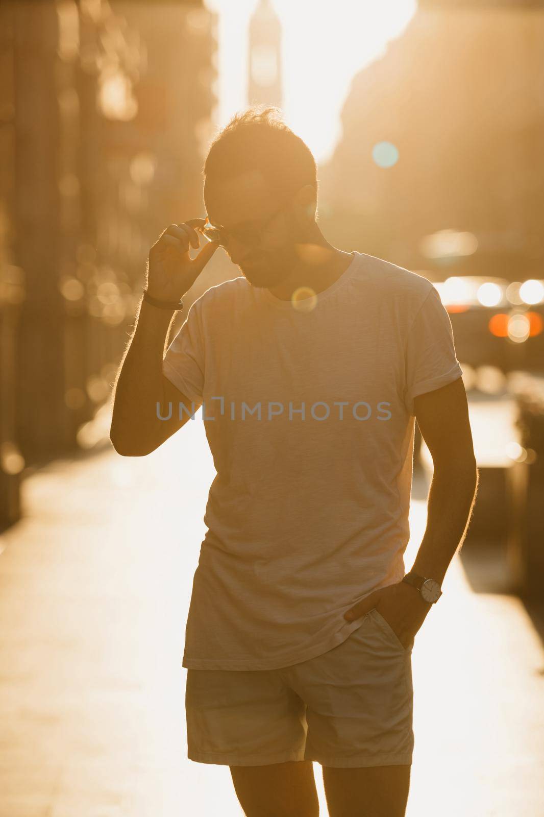 A man in a white t-shirt and shorts is straightening his sunglasses with his hand in the rays of the sunset on an old street in Spain. A guy with a beard is posing in Valencia in the evening.