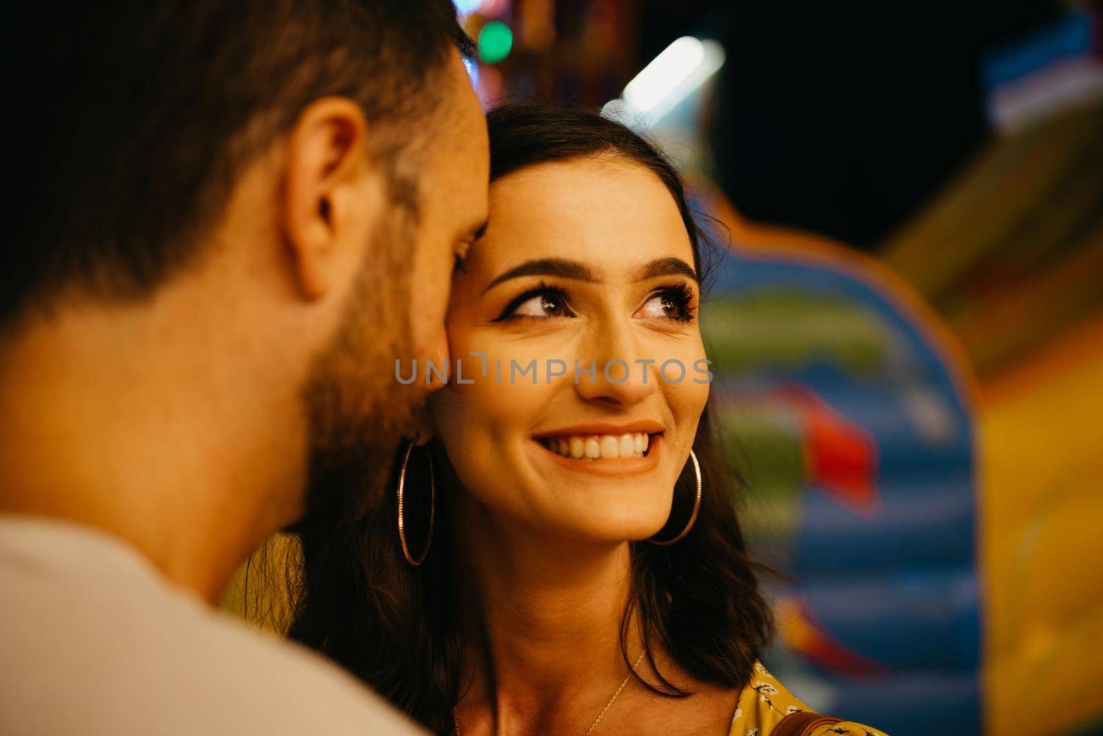 A close portrait of a woman in a yellow dress with a plunging neckline who is smiling to her boyfriend with a beard near amusement rides. A couple of lovers on a date at the fair in Valencia.