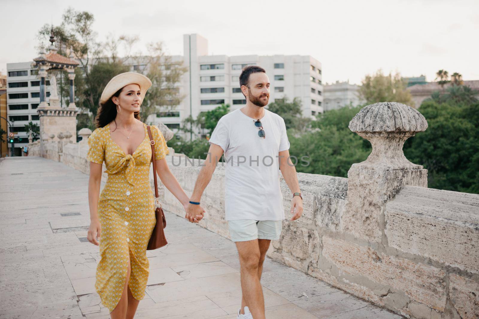 A girl in a hat and a yellow dress with a plunging neckline and her boyfriend with a beard are walking over the ancient bridge in old Spain town. A couple of tourists on a date in Valencia