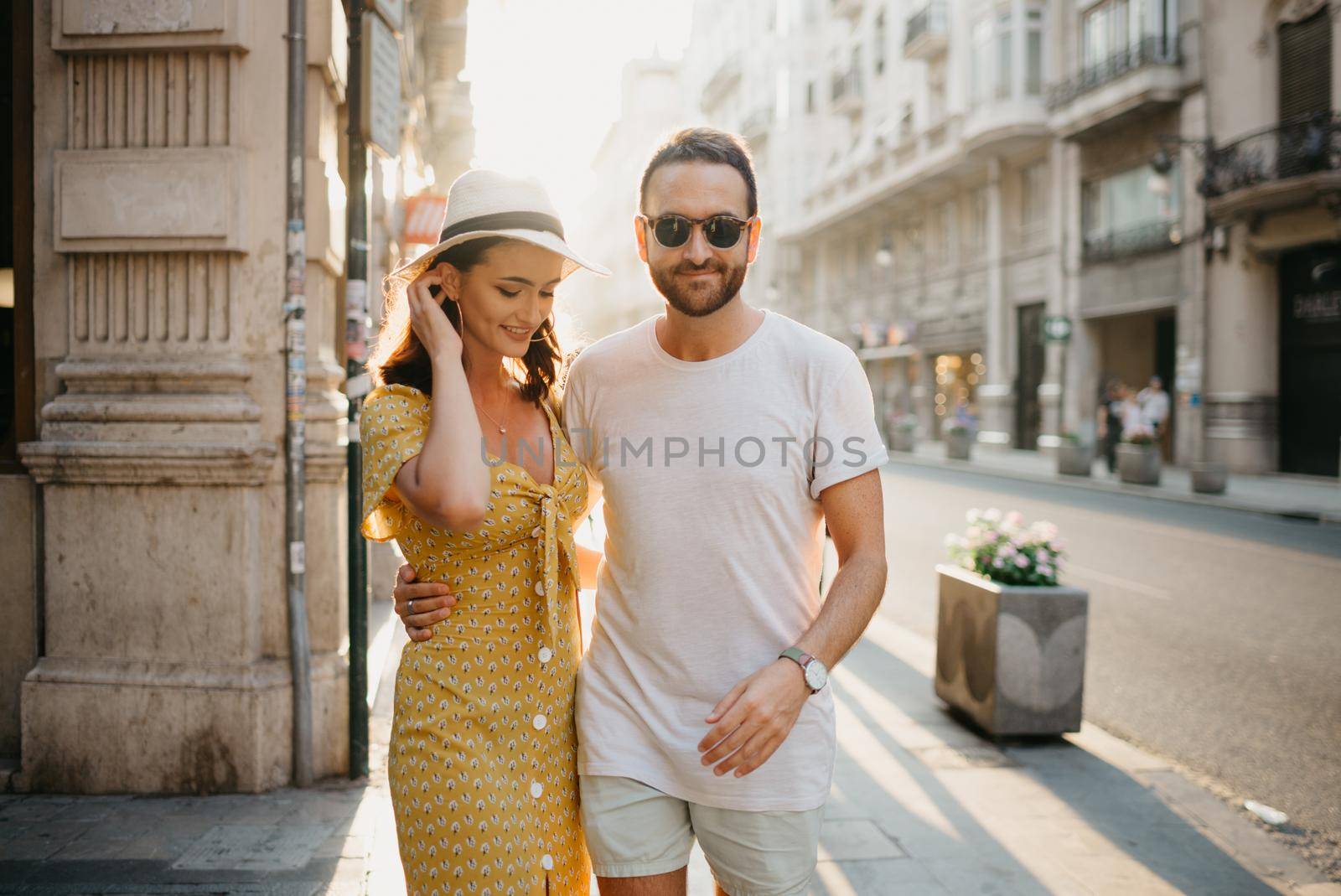 A man with a beard is hugging around the waist with his hand of a girlfriend in a hat and a yellow dress with a plunging neckline in Spain. A couple of lovers on a date on the sunset in Valencia.