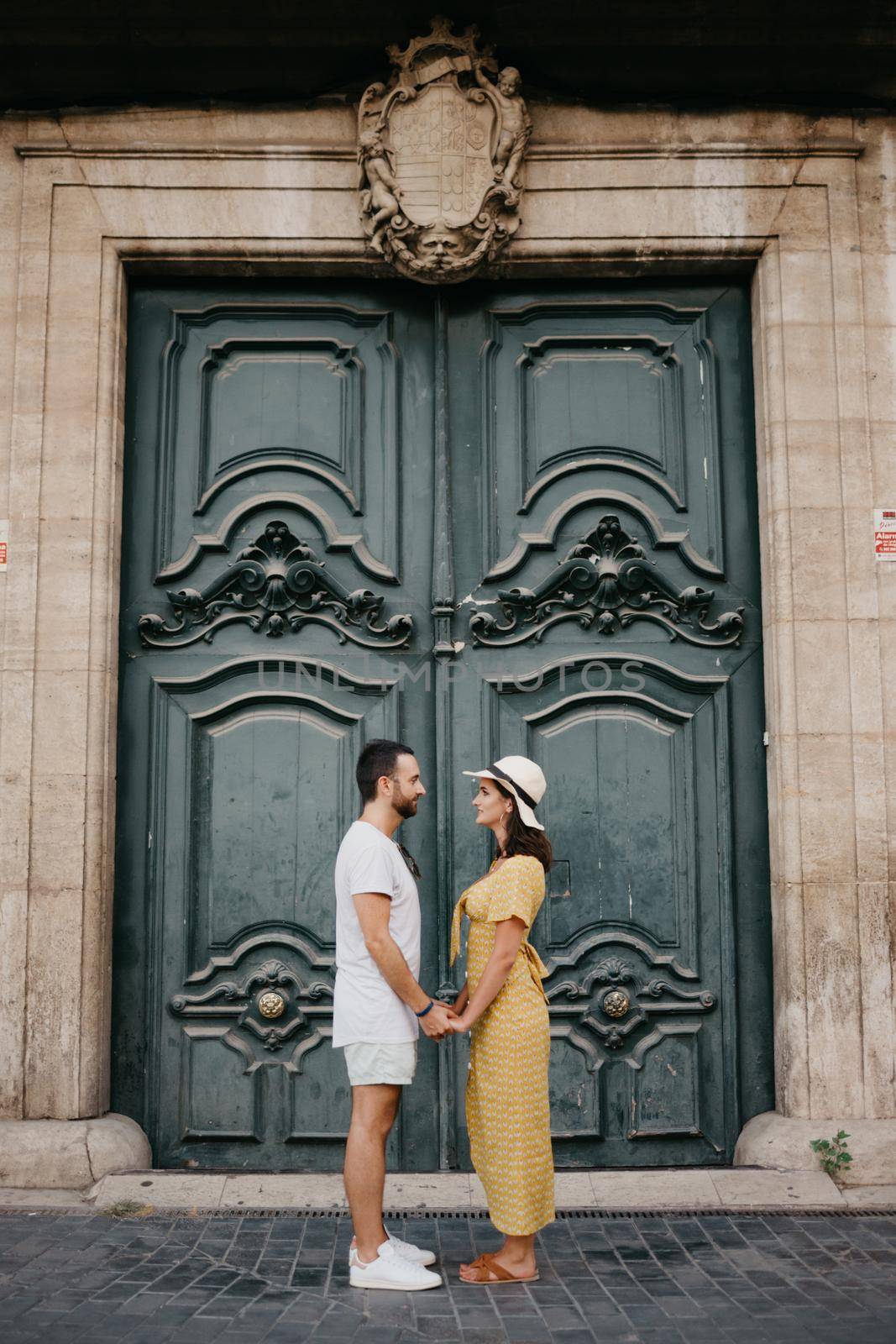 A girl in a hat and a dress with a plunging neckline and her boyfriend with a beard are posing near the giant doors holding each other's hand in the old town. A couple of tourists in Valencia.