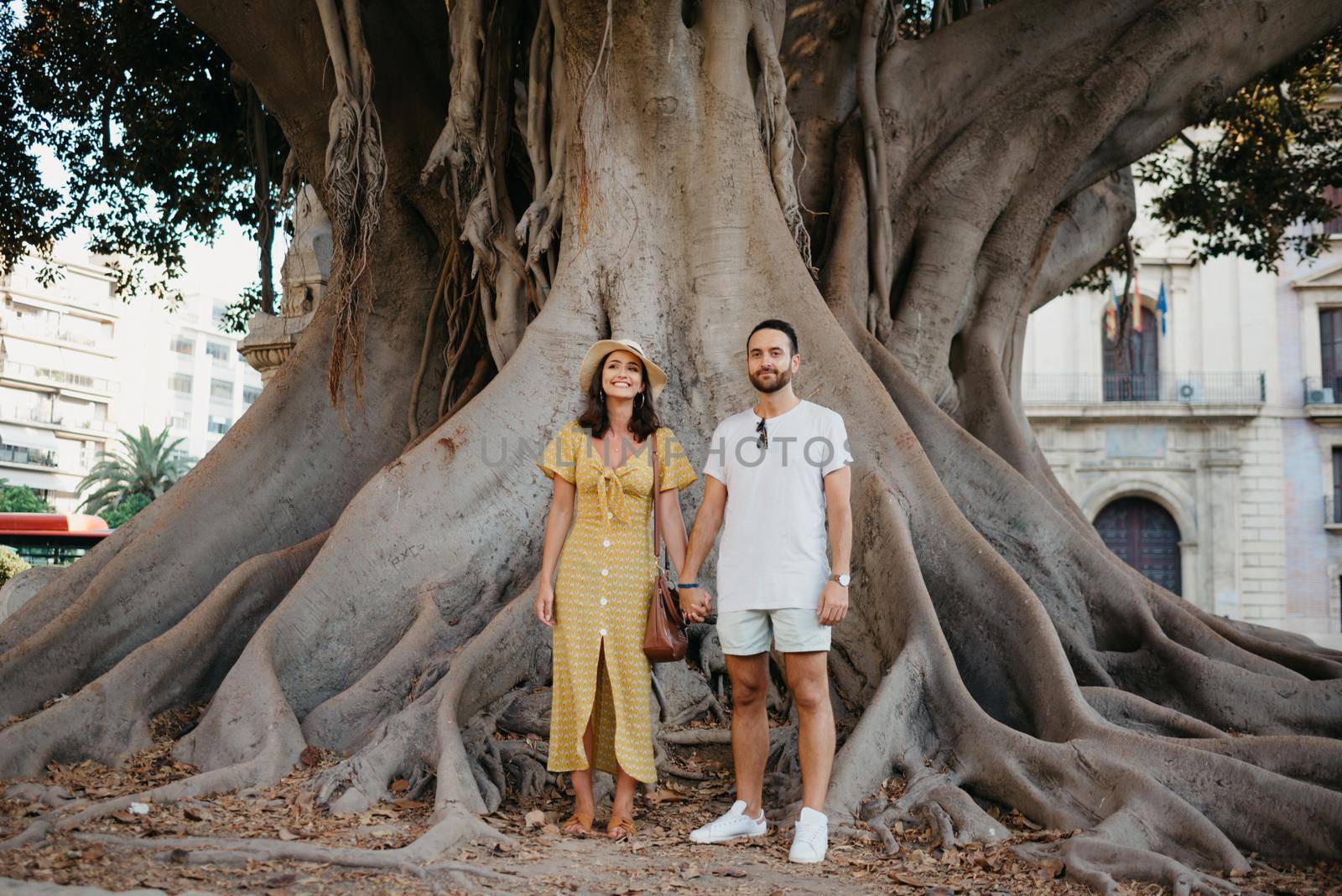 A beautiful young brunette in a hat and yellow dress with her boyfriend with a beard under an old giant Valencian Ficus Macrophylla tree in Spain in the evening. A couple of tourists in Valencia.