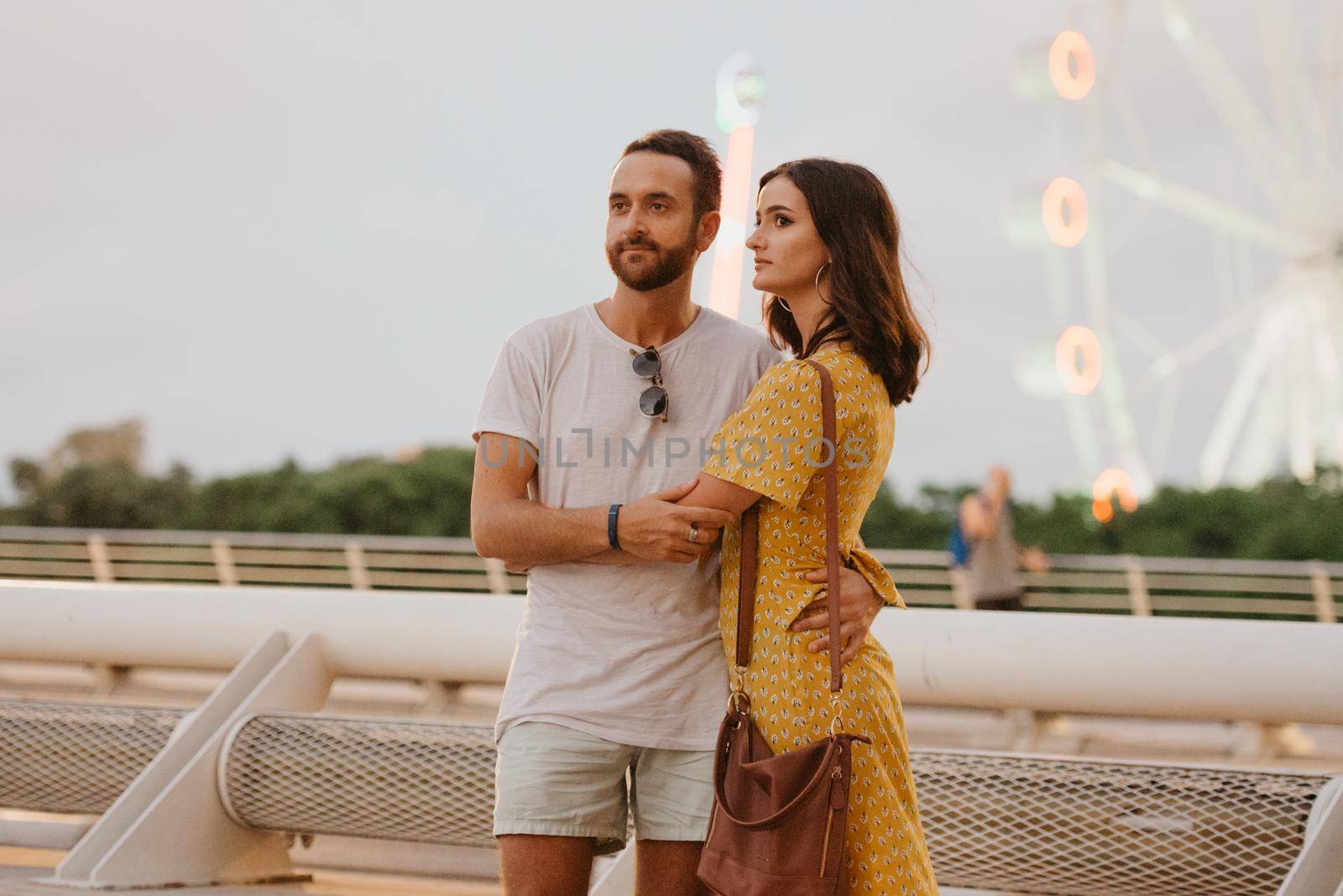 A brunette girl in a yellow dress and her boyfriend are hugging on a white bridge with a Ferris wheel in the background in Valencia. A couple of tourists on a date in the warm evening.