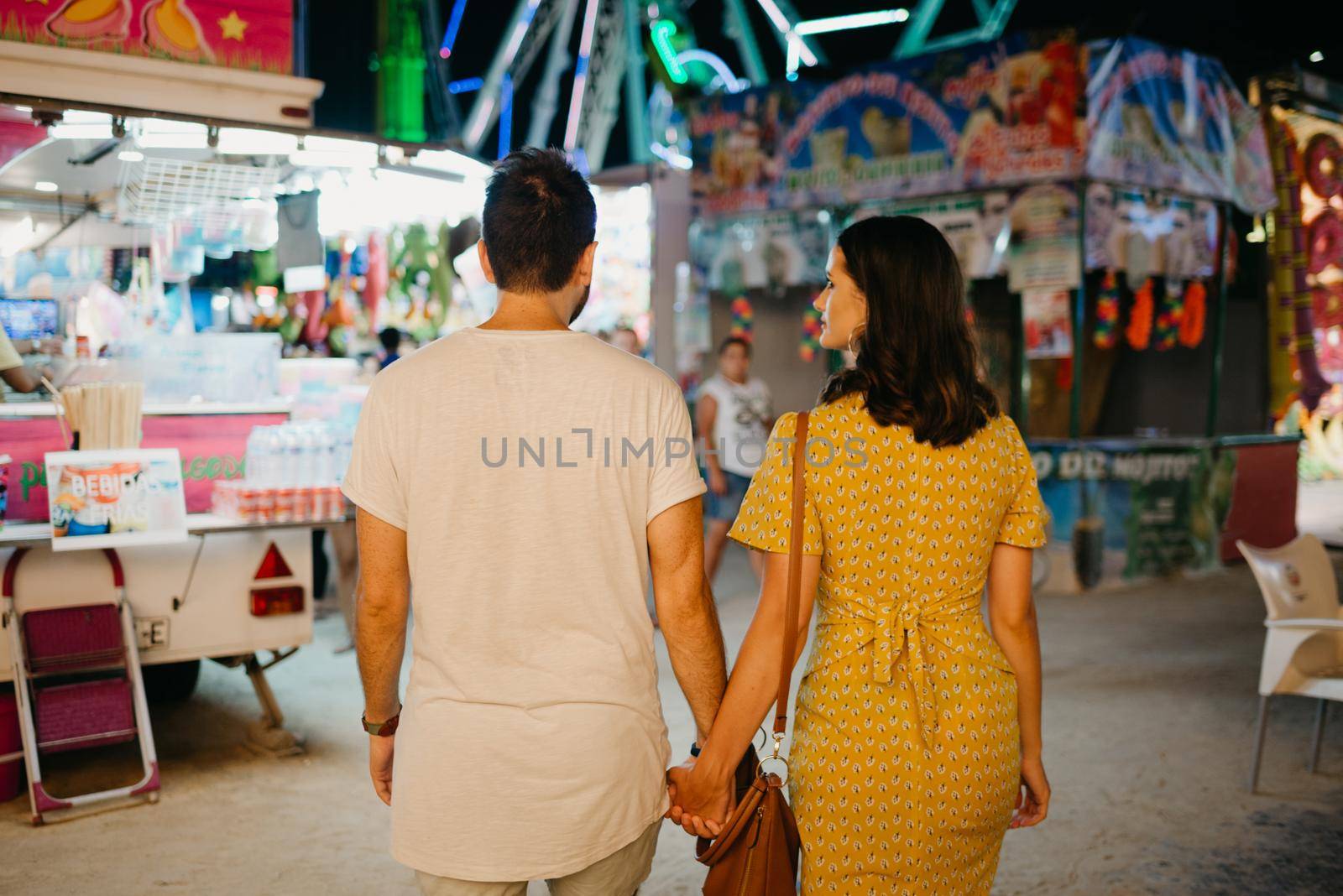 A girl in a yellow dress and her boyfriend with their backs are walking between amusement rides. A couple of lovers on a date at the fair in Valencia.