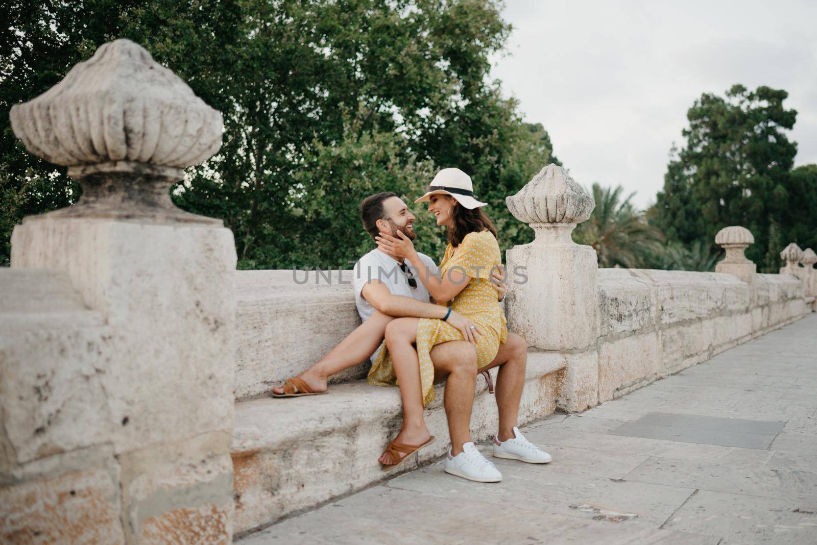 A girl in a hat and a yellow dress with a plunging neckline is sitting on the legs of her boyfriend with a beard on the ancient bridge in old Spain town. A couple of tourists on a date in Valencia.