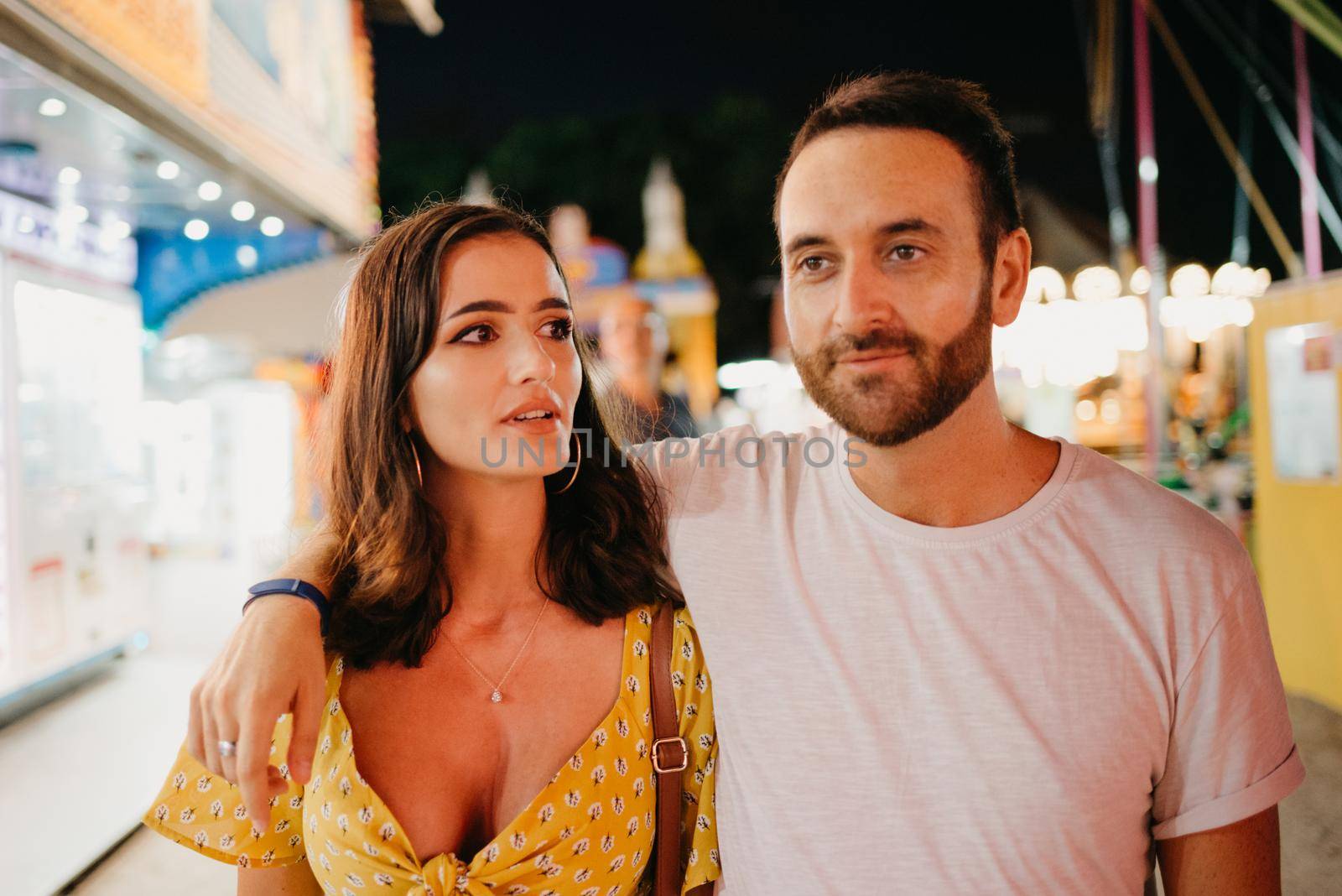 A close portrait of a girl in a yellow dress and her boyfriend with a beard are hugging each other and walking between amusement rides. A couple of lovers on a date at the fair in Valencia.