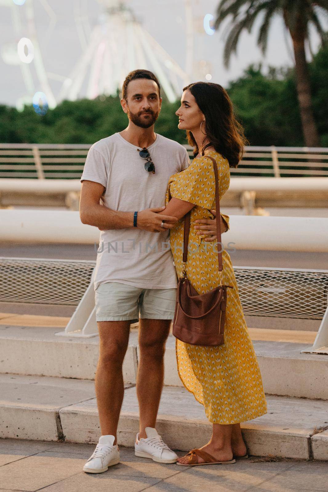 A brunette girl in a yellow dress and her boyfriend are hugging on a white bridge with a Ferris wheel in the background in Valencia. A couple of tourists in a full length on a date in the warm evening