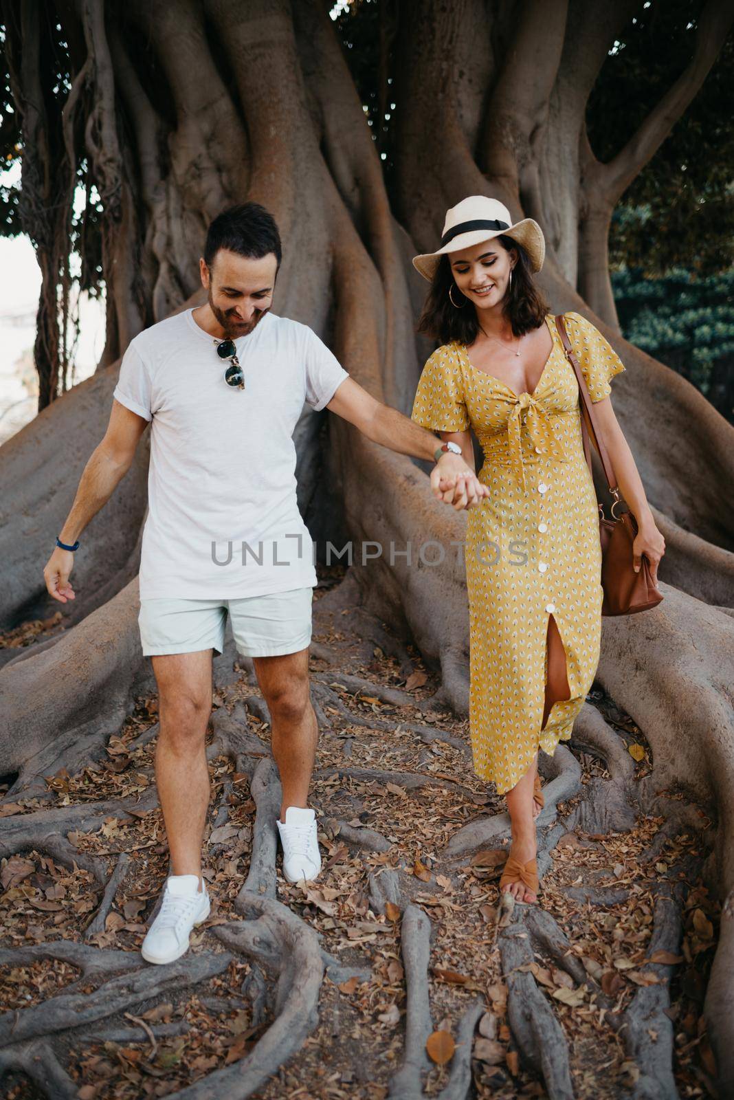 A young woman in a hat and a yellow dress and her boyfriend with a beard walk under an old Valencian Ficus Macrophylla in Spain. A couple of lovers are exploring Valencia in the evening.