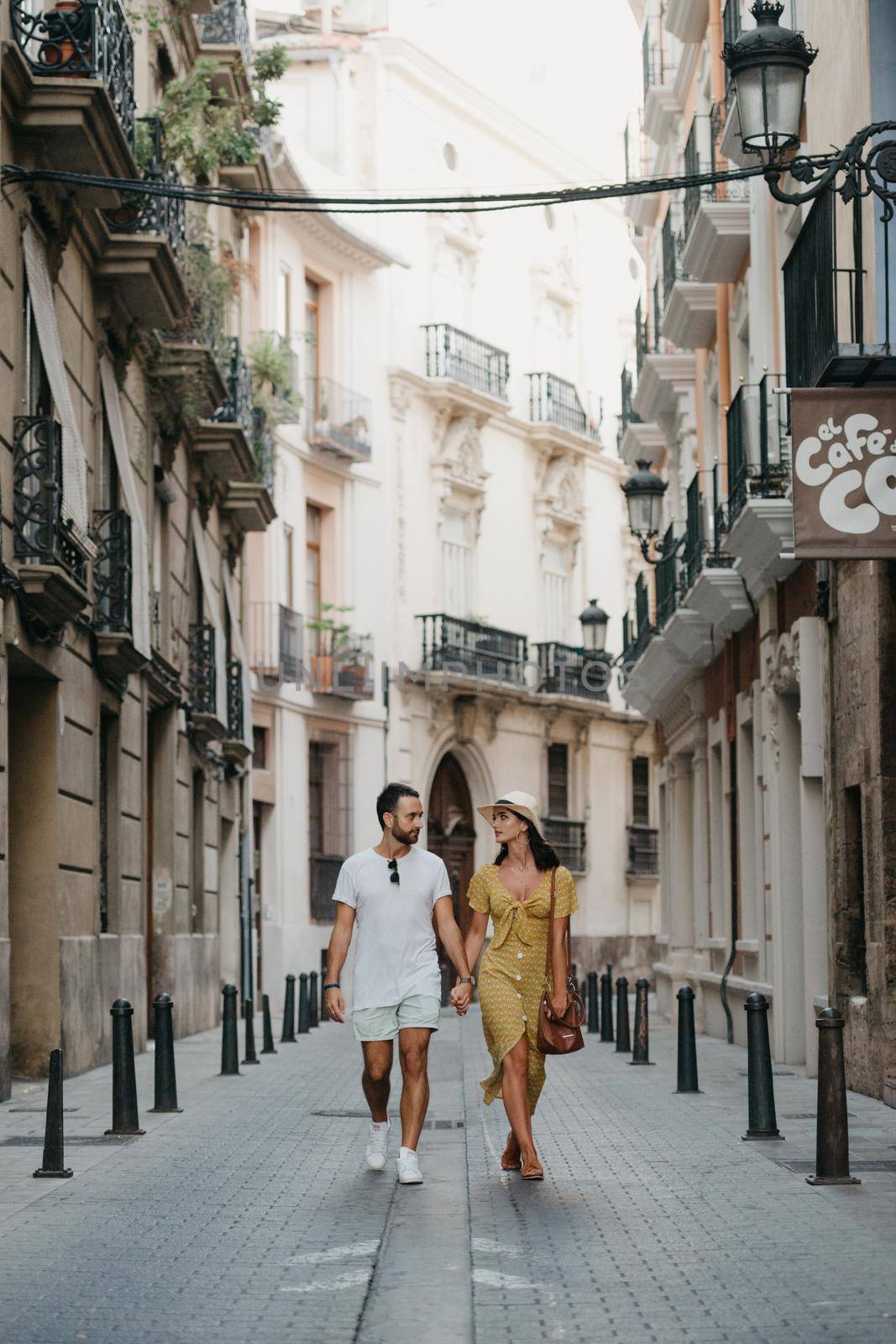 A girl in a hat and a yellow dress with a plunging neckline and her boyfriend with a beard are walking and staring at each other in old Spain town. A couple of tourists on a date in Valencia.