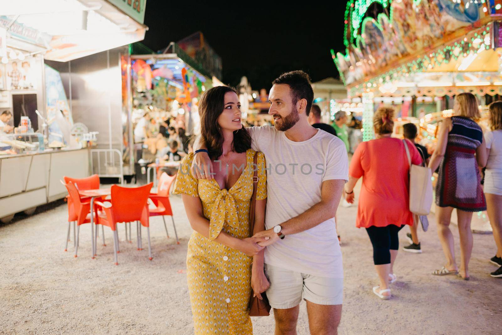 A young woman in a yellow dress and her boyfriend with a beard are hugging each other and walking between amusement rides. A couple of lovers on a date at the fair in Valencia.