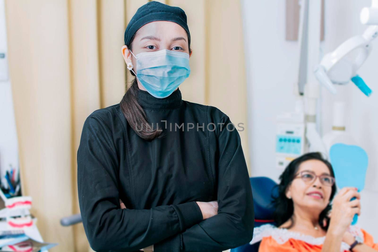 Dental doctor standing in clinic wearing mask, portrait of dentist crossing arms with patient in background, Modern Dental clinic. Dental procedures. portrait of a dentist woman with crossed arms