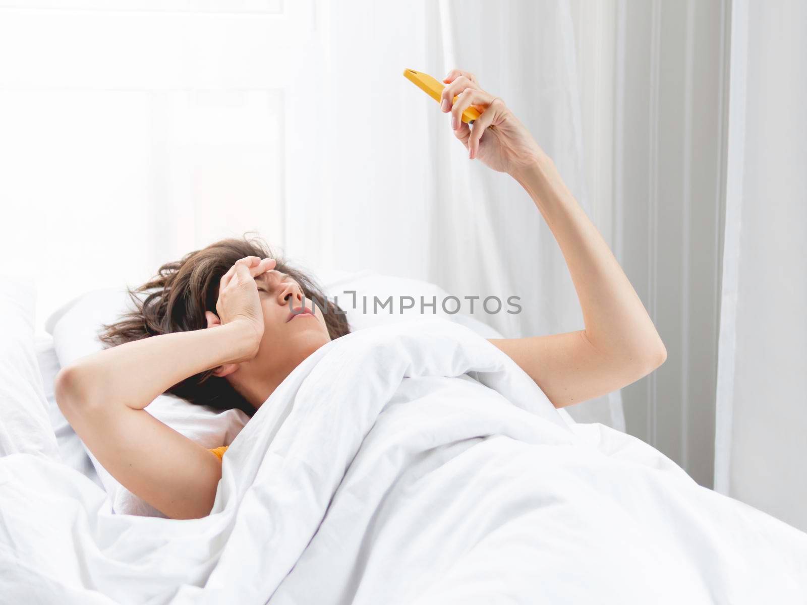 Sleepy woman looks on smartphone screen. Checking e-mail box right after waking up. Using wireless technology in bed. by aksenovko