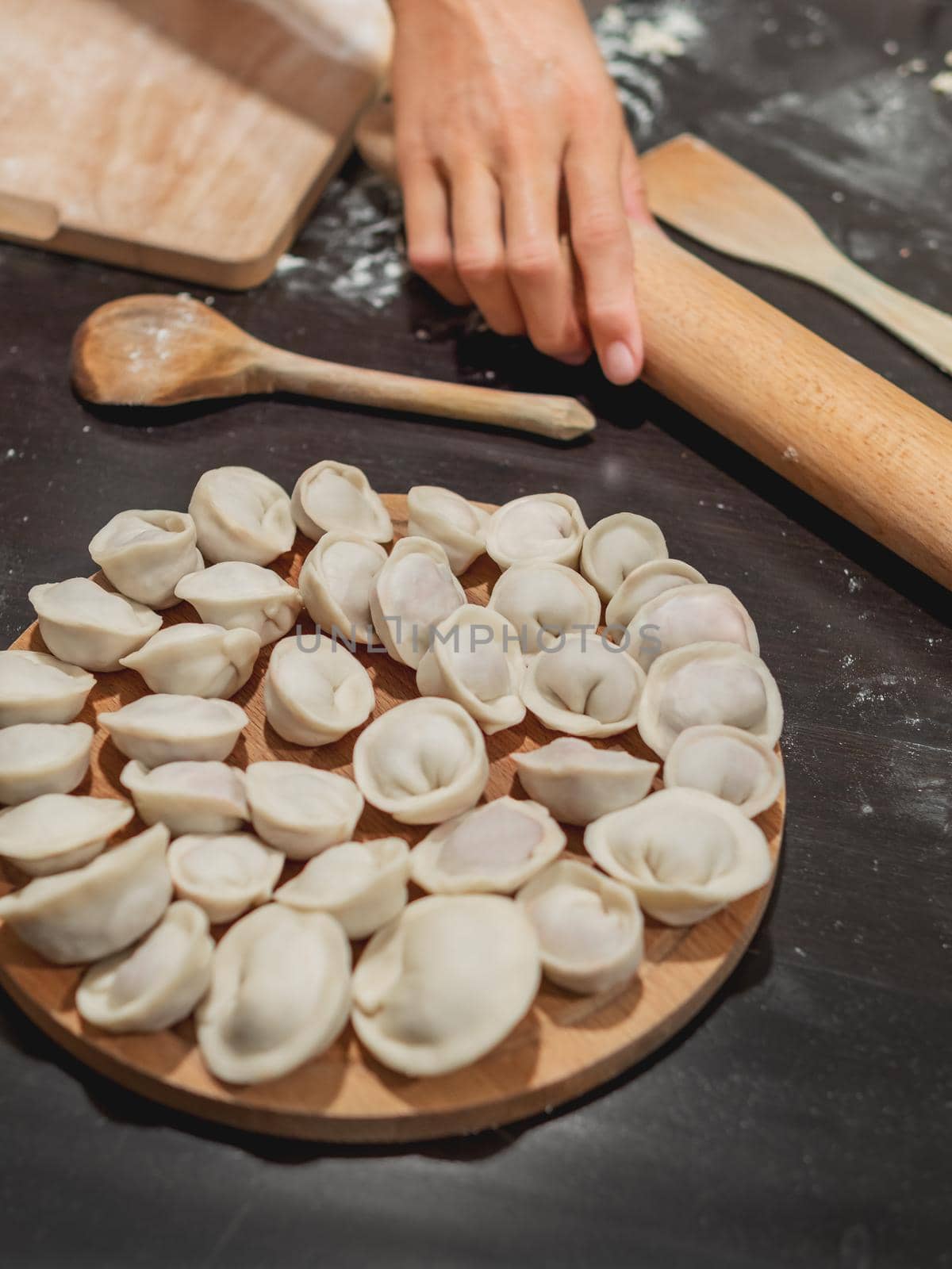 Woman makes pelmeni or dumplings - traditional dish of Russian cuisine made of dough and meat. Black kitchen table with flour and wooden utensils.