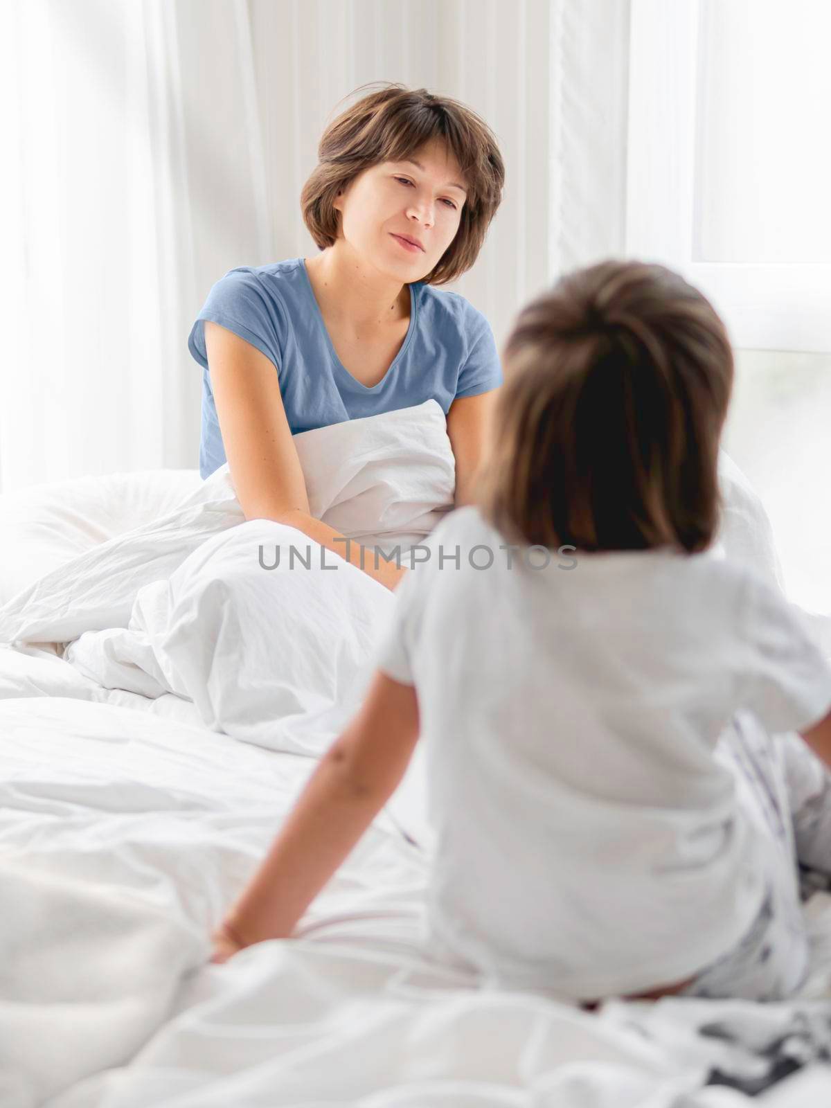 Kid wakes his mother up. Sleepy disheveled mom sits in bed. Uneasy life as parent. Bedroom lit with morning light. by aksenovko