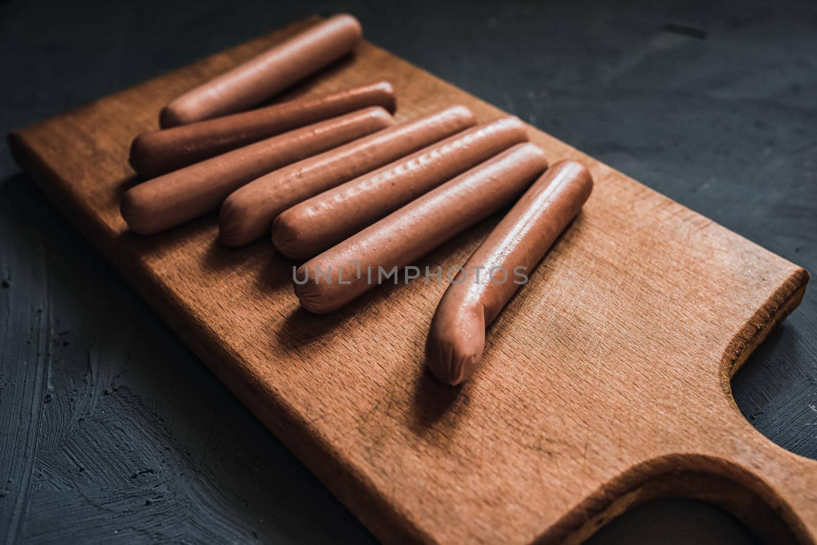 Boiled fried sausages sausages lie on a wooden kitchen board scratched against a dark concrete background.