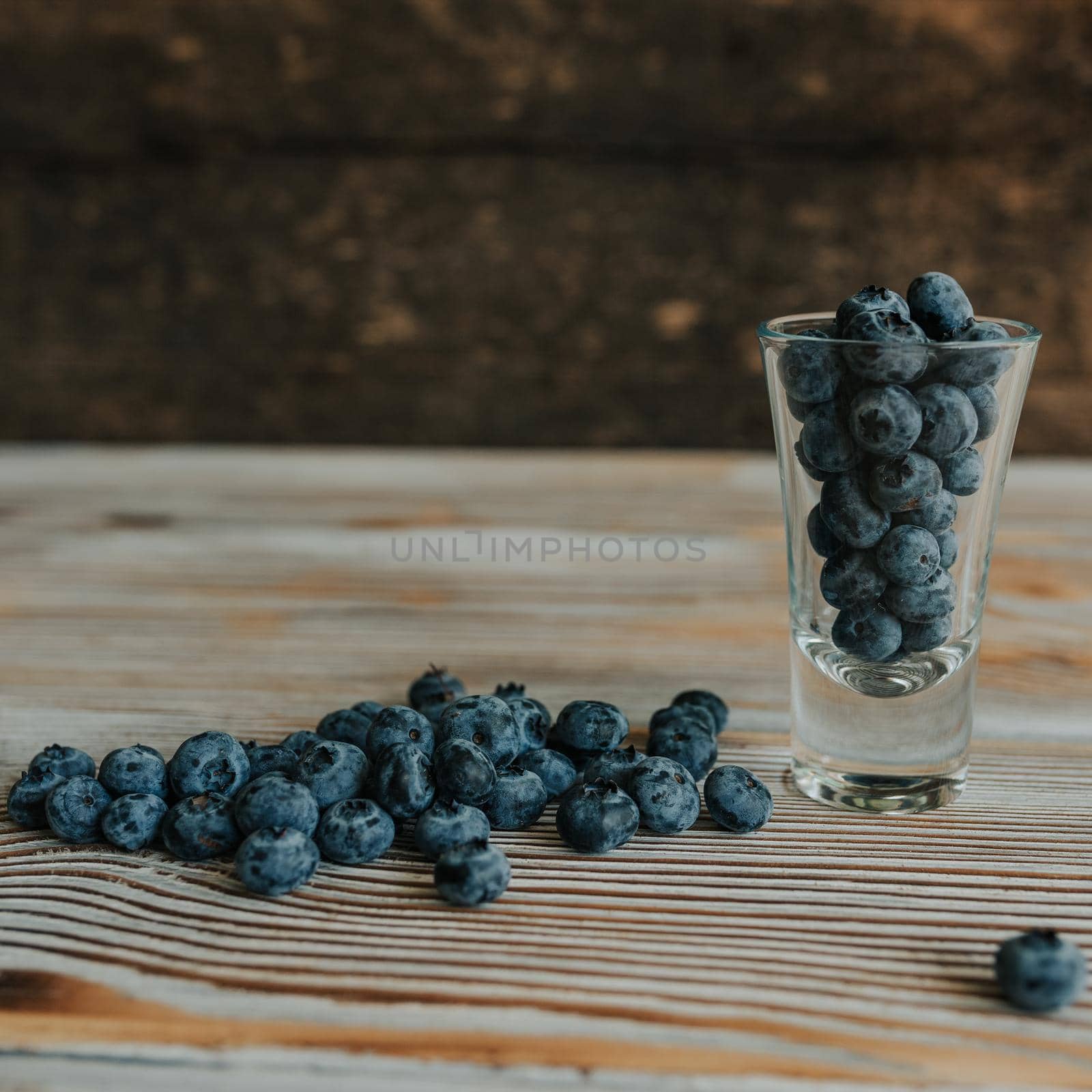 Fresh blue matte round blueberries filled in a transparent glass stands on a wooden brown shabby textured table and berries are scattered everywhere
