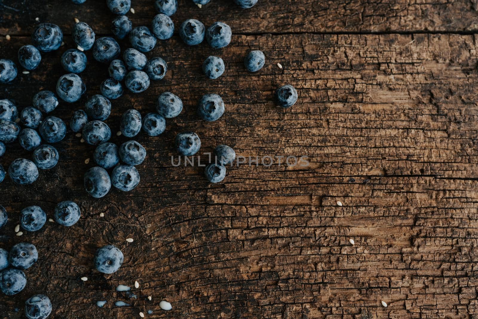 Blue fresh blueberries are scattered on an old brown wooden cracked table. Sesame seeds were poured between the fruits. Wet drops of milk on the table. basil leaves, green fresh seasoning.