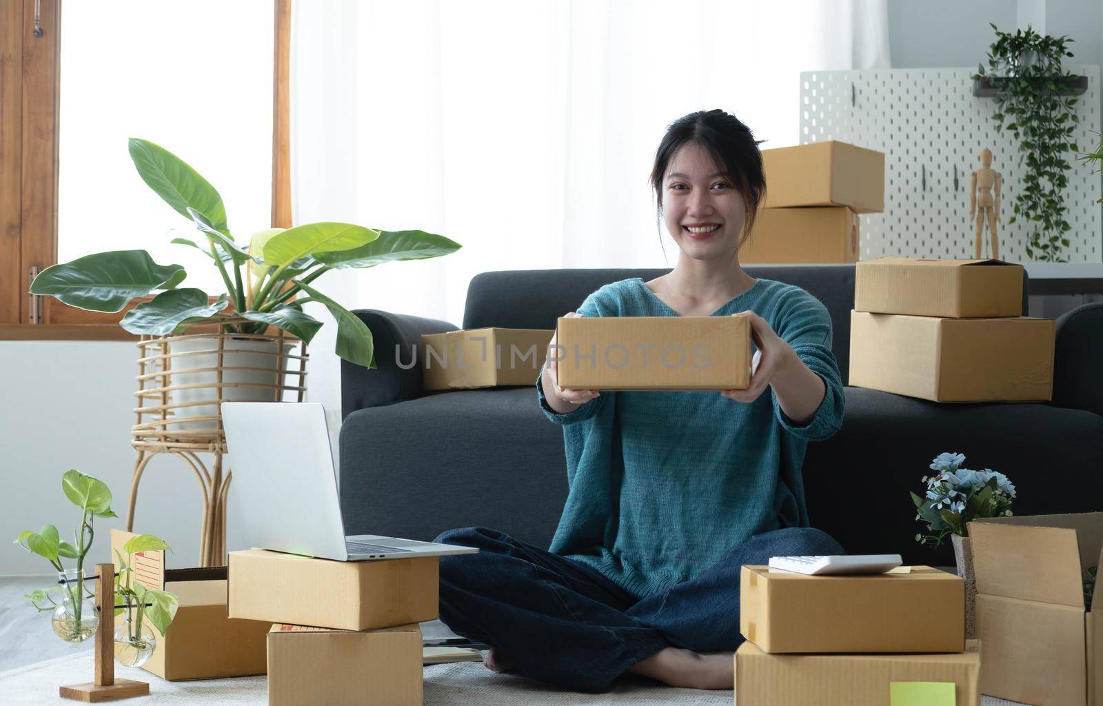 Happy young asian woman startup small business freelance holding parcel box and computer laptop and sitting on floor, Online marketing packing box delivery concept by wichayada