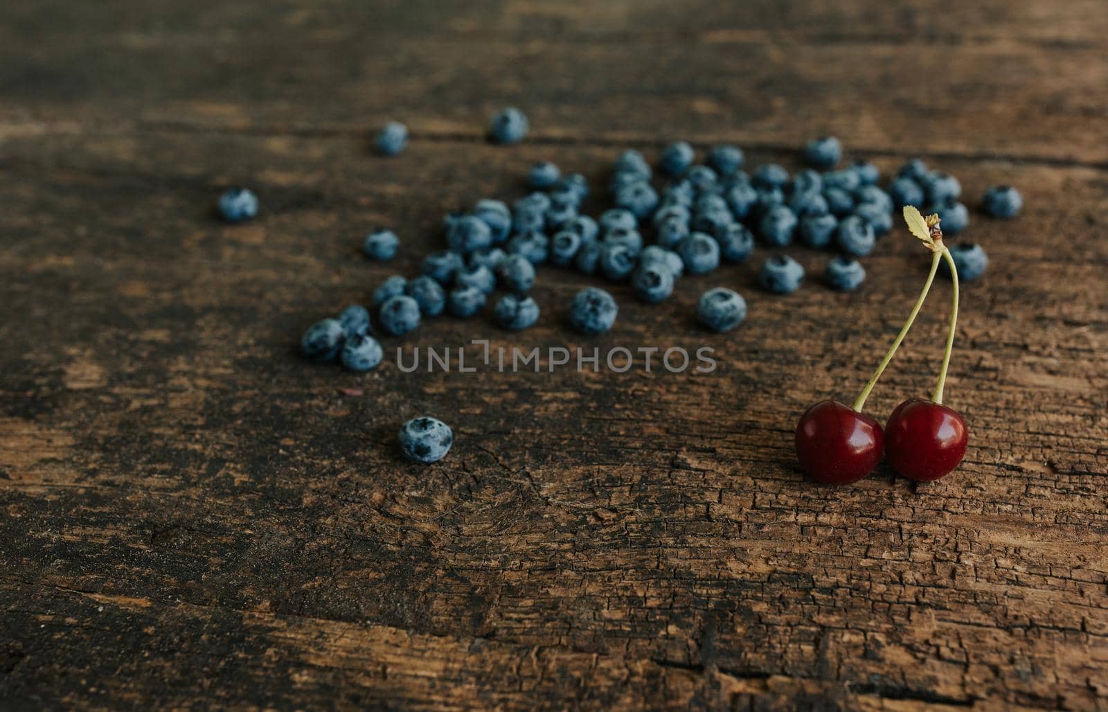 Blue fresh blueberries are scattered on an old brown wooden cracked table.Two red cherries with roots stand on a wooden background.