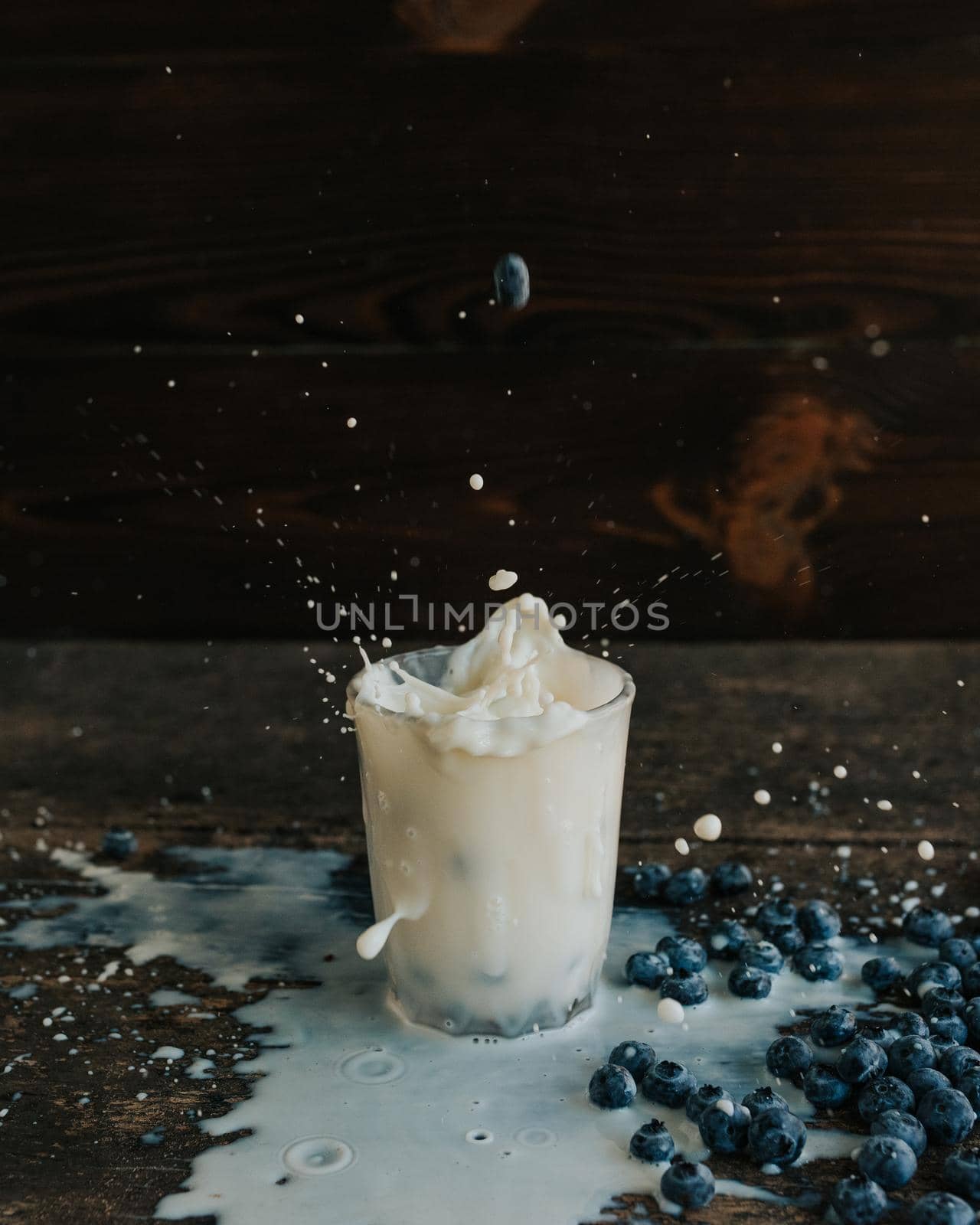 Berries are thrown into the transparent glass making splashes of milk. Blue fresh blueberries are scattered on an old brown wooden cracked table. White water pour down on the table.