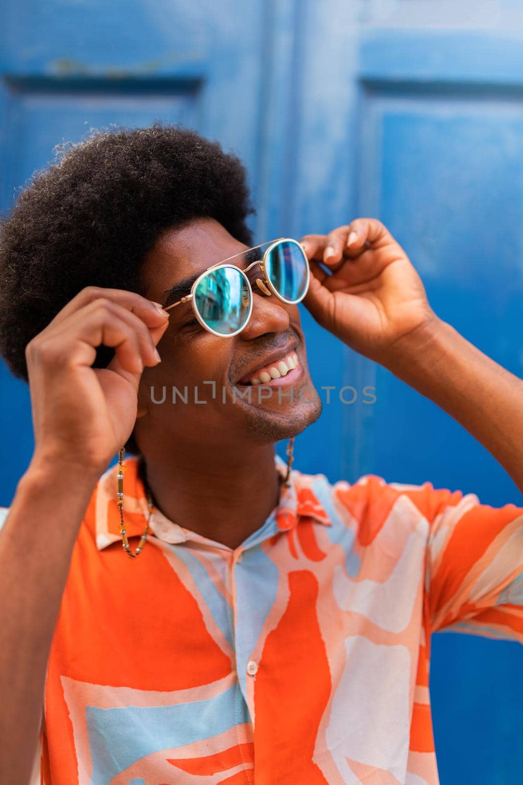 Vertical portrait of young happy black man putting on sunglasses outdoors. by Hoverstock