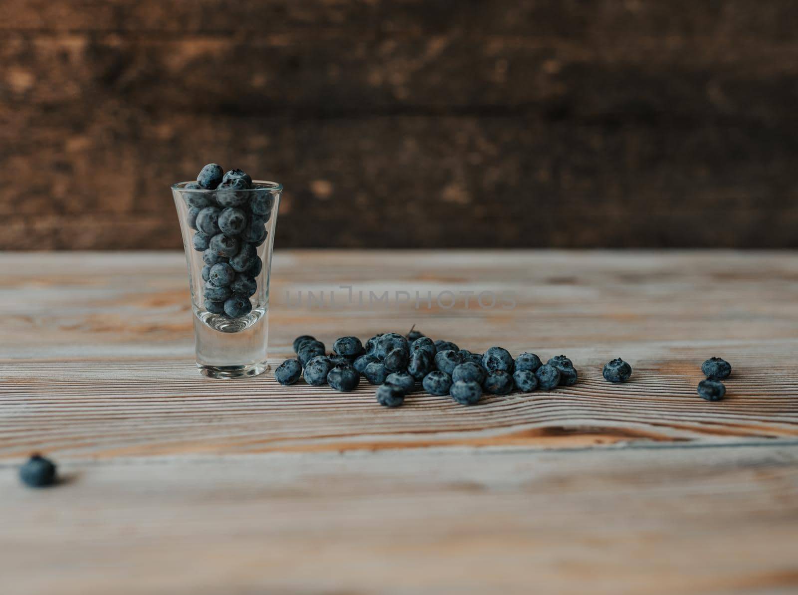 Fresh blue matte round blueberries filled in a transparent glass stands on a wooden brown shabby textured table and berries are scattered everywhere