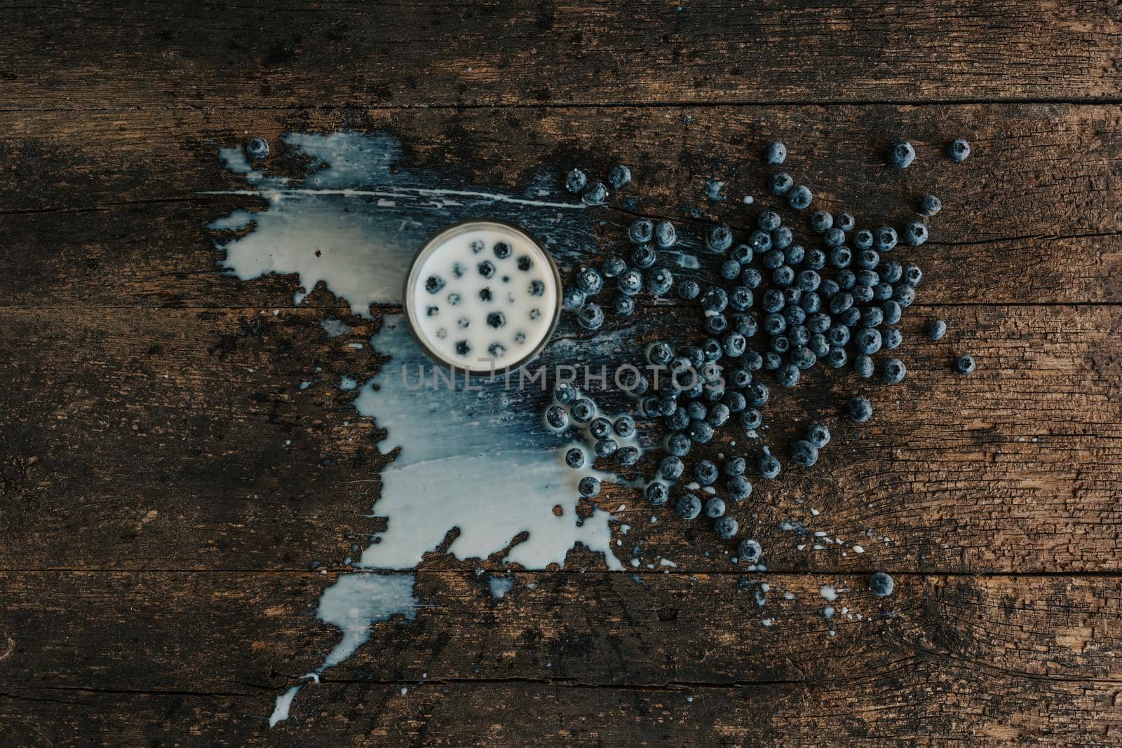 Berries are thrown into the transparent glass making splashes of milk. Blue fresh blueberries are scattered on an old brown wooden cracked table. White water pour down on the table.